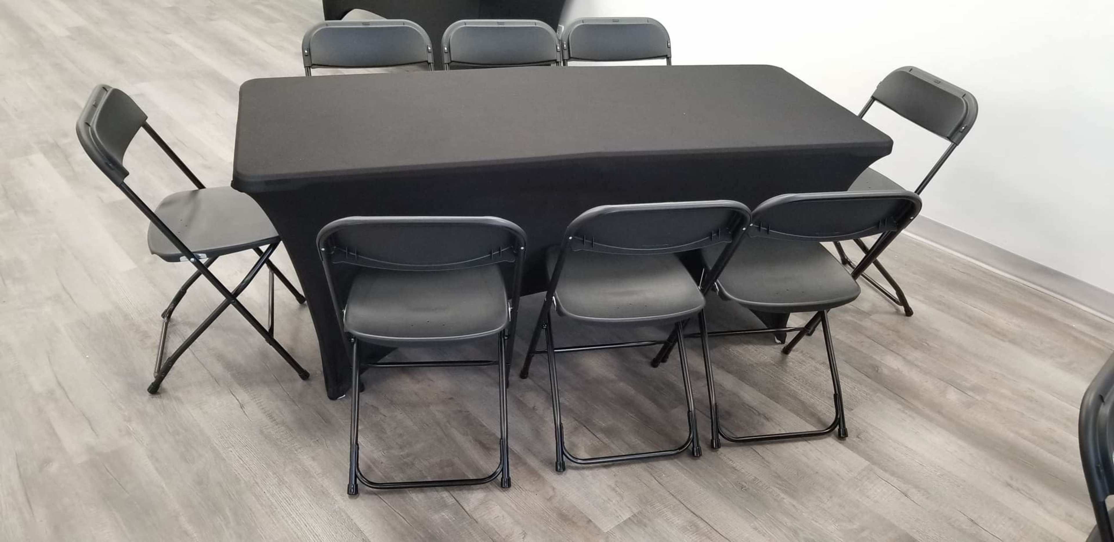 A rectangular table with a black cover is surrounded by six black folding chairs in a room with wooden flooring.