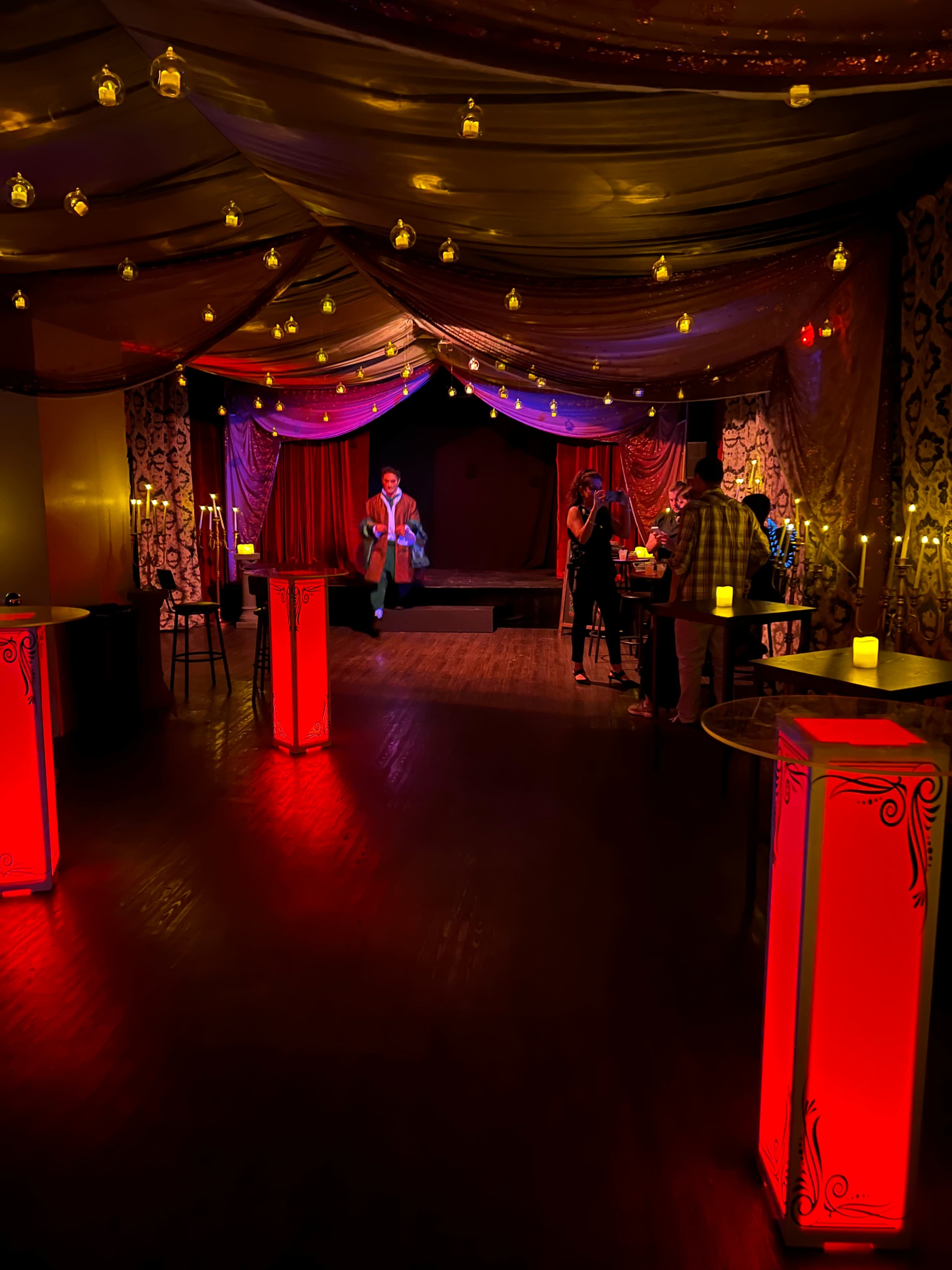 The image shows a dimly lit lounge with red illuminated tables, draped fabric overhead, and people mingling in front of a small stage.