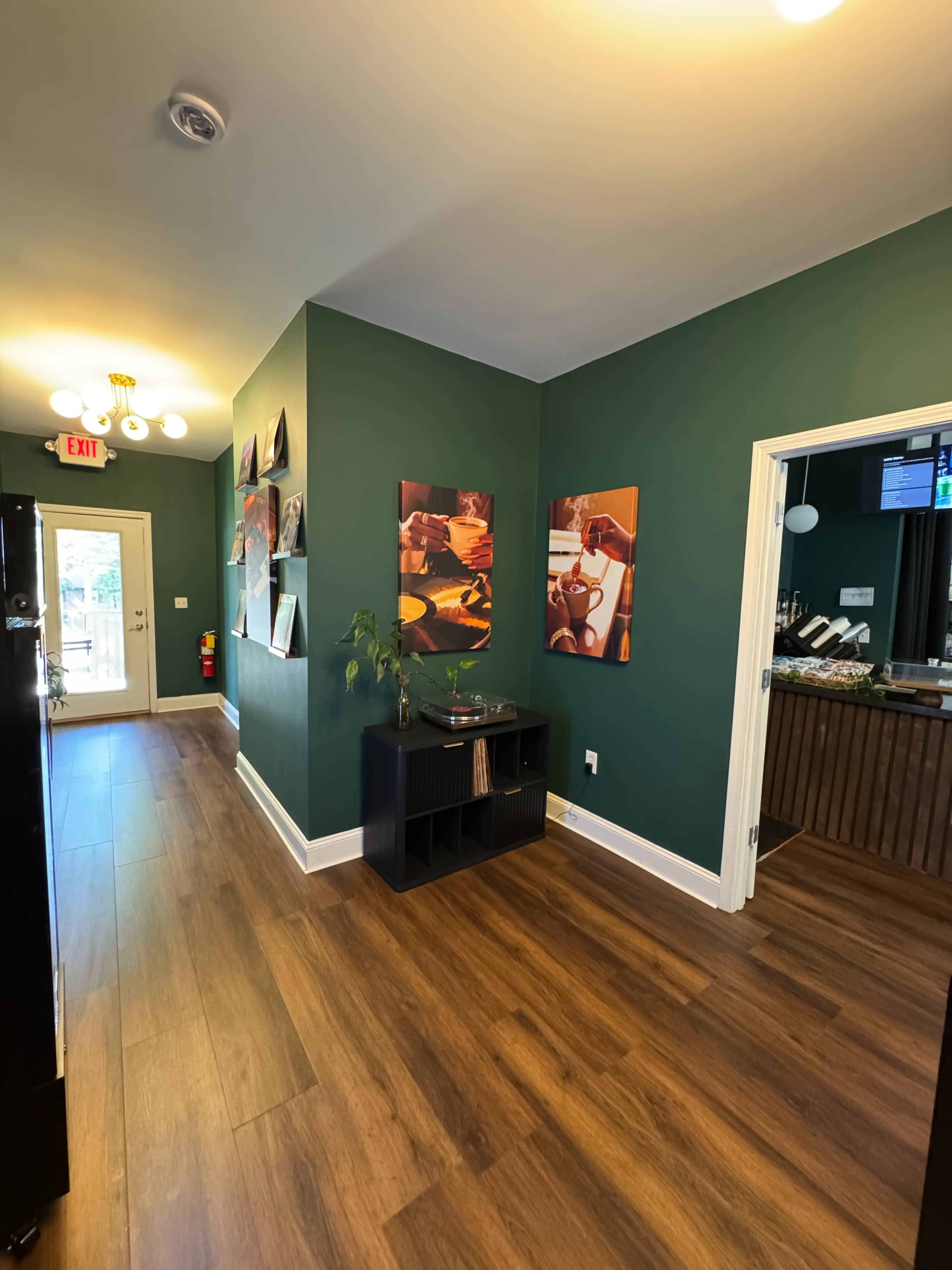 The image shows a hallway with green walls, wooden flooring, and framed artwork, leading to an exit door and an adjoining room with a bar counter.