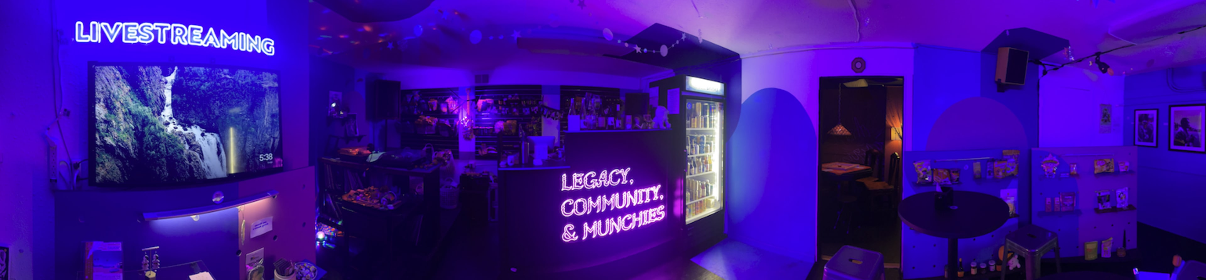 The image shows a dimly lit, modern lounge or café with purple lighting, featuring a bar area with snacks, drinks, and a screen displaying a nature scene.