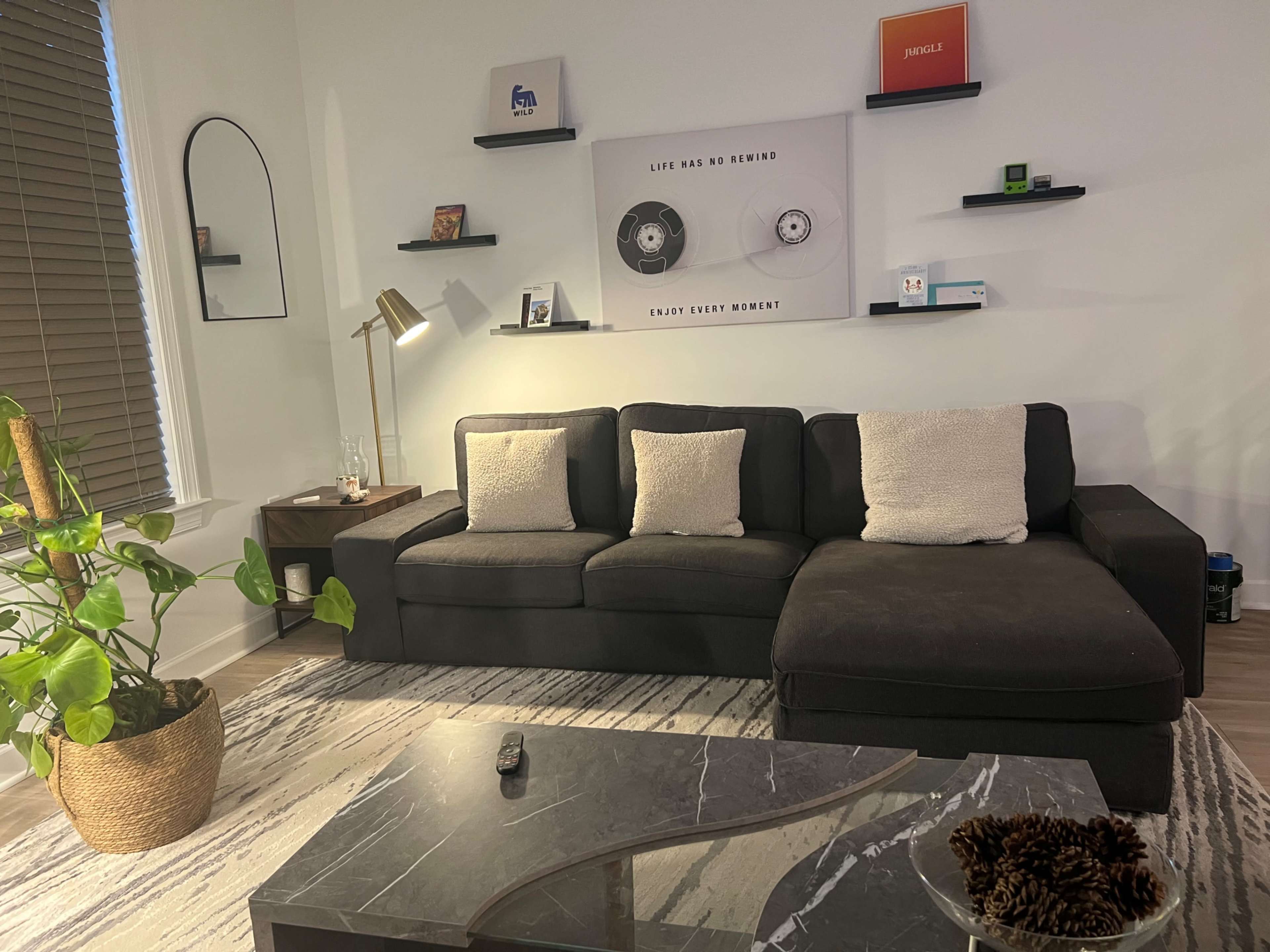 The image shows a modern living room featuring a black sectional sofa with four pillows, a coffee table, and decorative shelves on the wall above.
