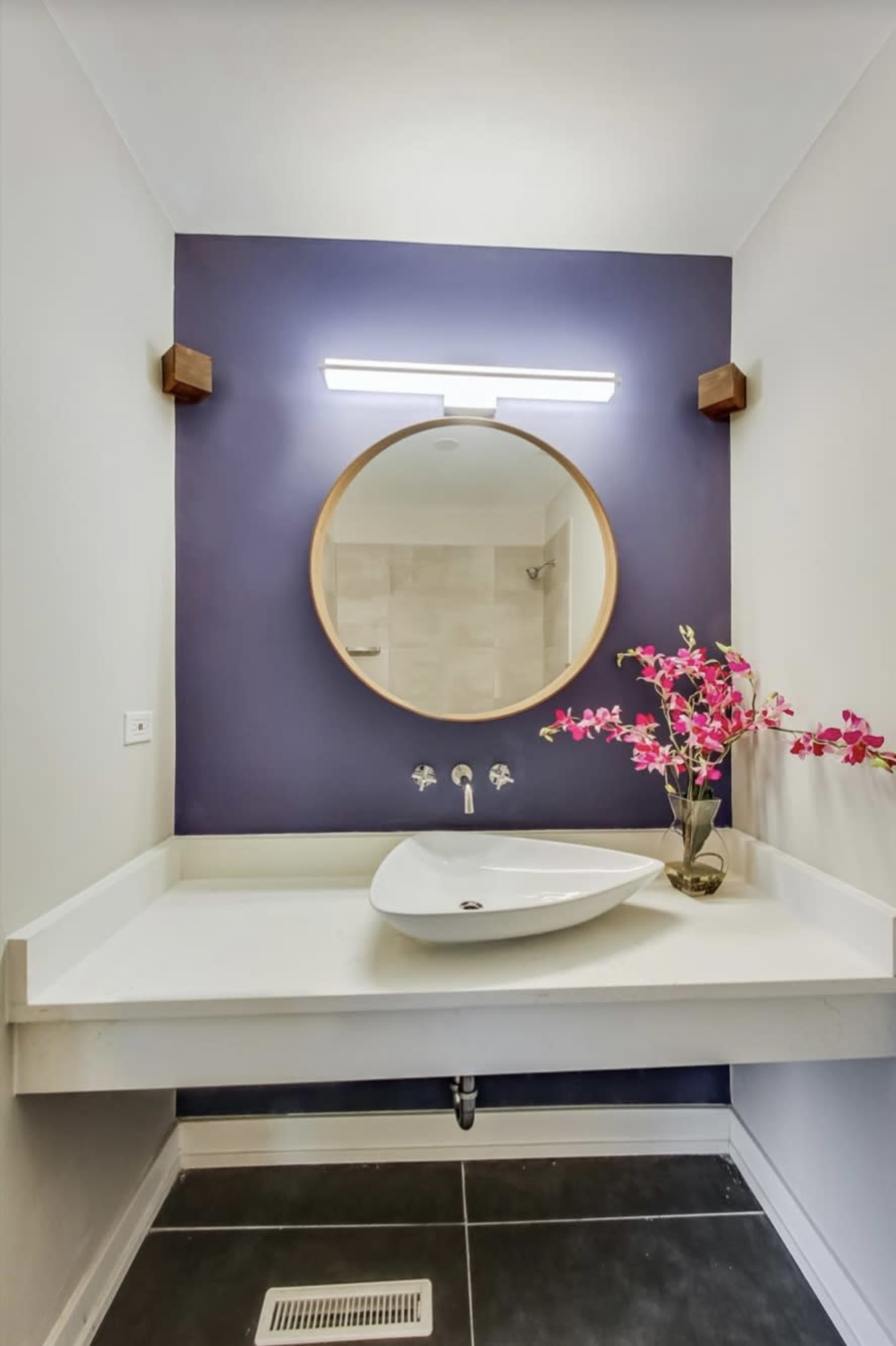 A modern bathroom with a circular mirror, a wall-mounted light fixture, a white sink, and a decorative vase with flowers on a counter against a dark purple wall.