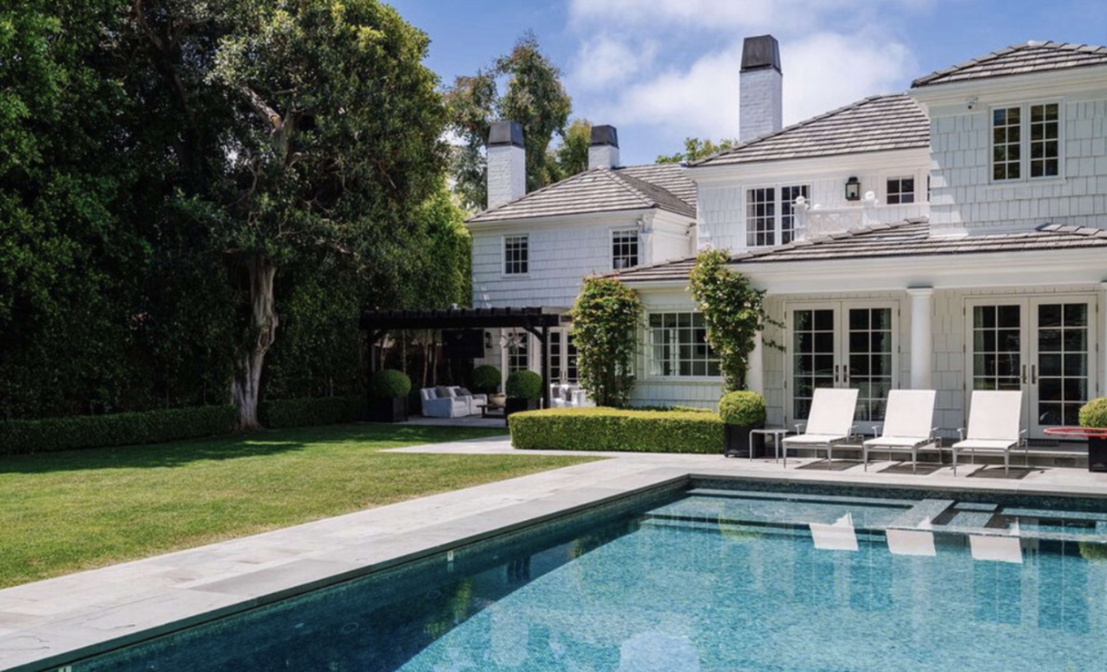 The image shows a spacious backyard featuring a rectangular swimming pool, lounge chairs, and a well-maintained lawn surrounded by a white two-story house with a patio area.