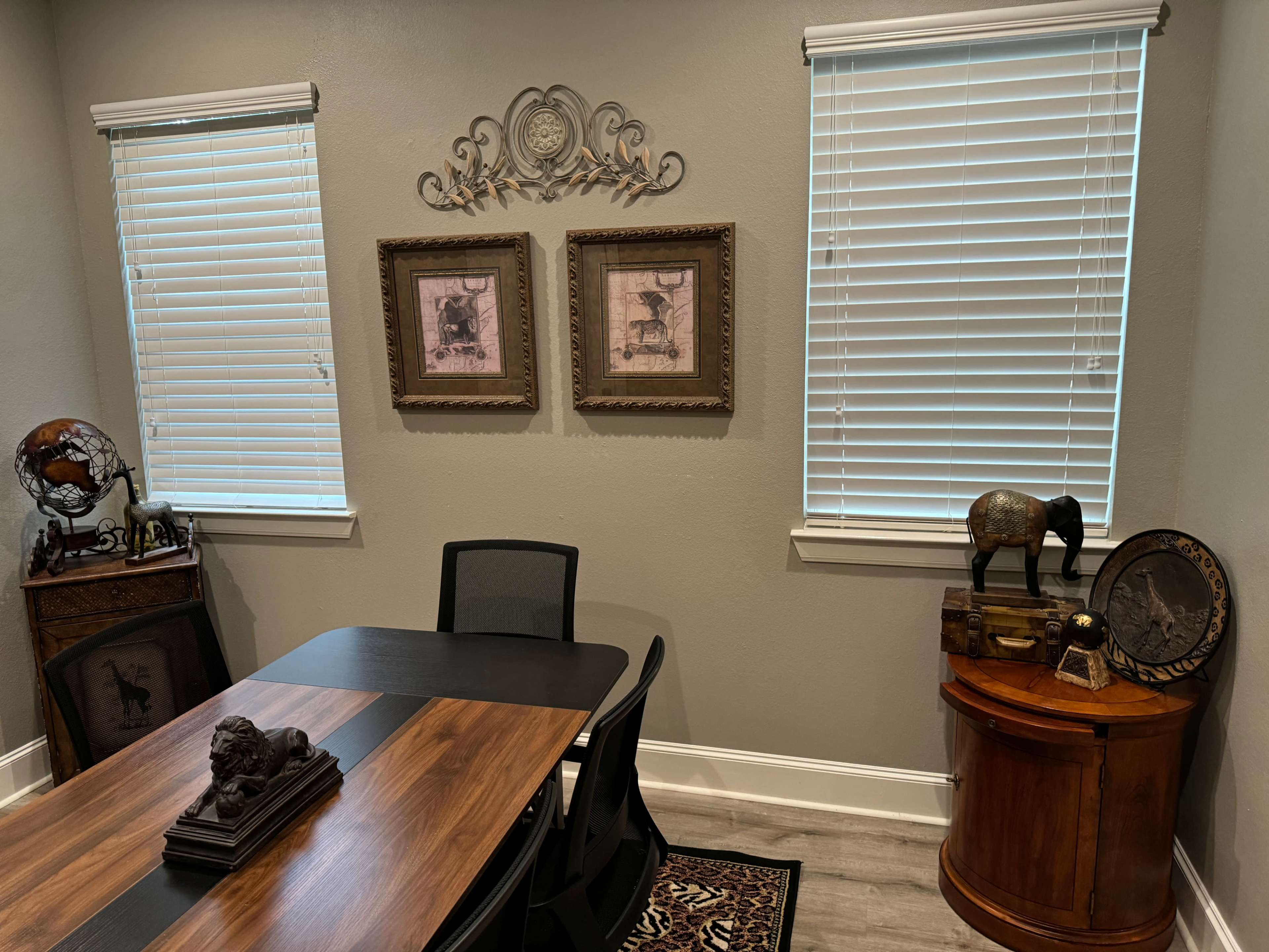 The image shows a neatly arranged room with a table in the center, framed artwork on the walls, and decorative elements in the corners.
