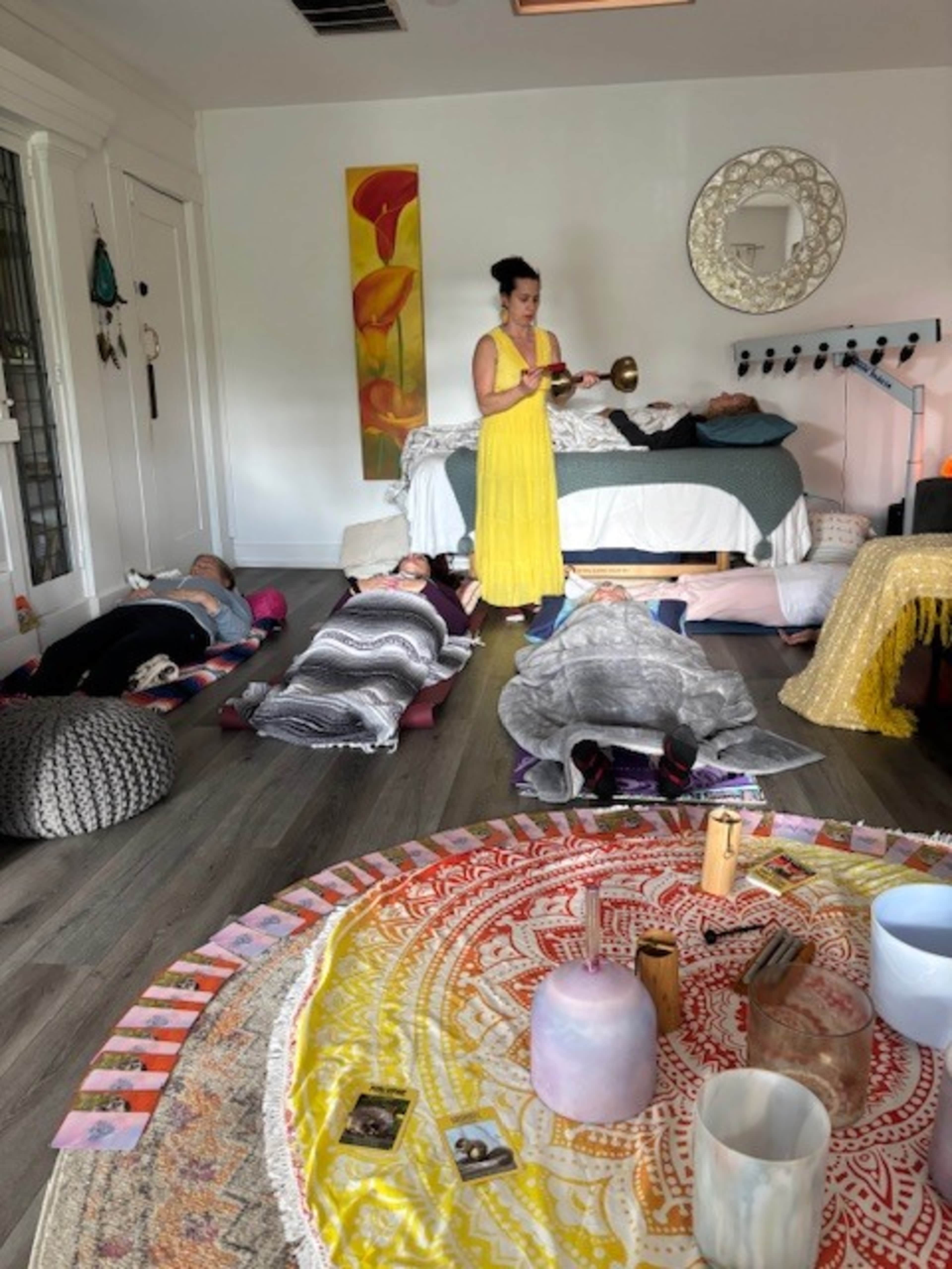 A woman in a yellow dress stands in a room while several people lay on the floor, surrounded by various items and decorative elements.