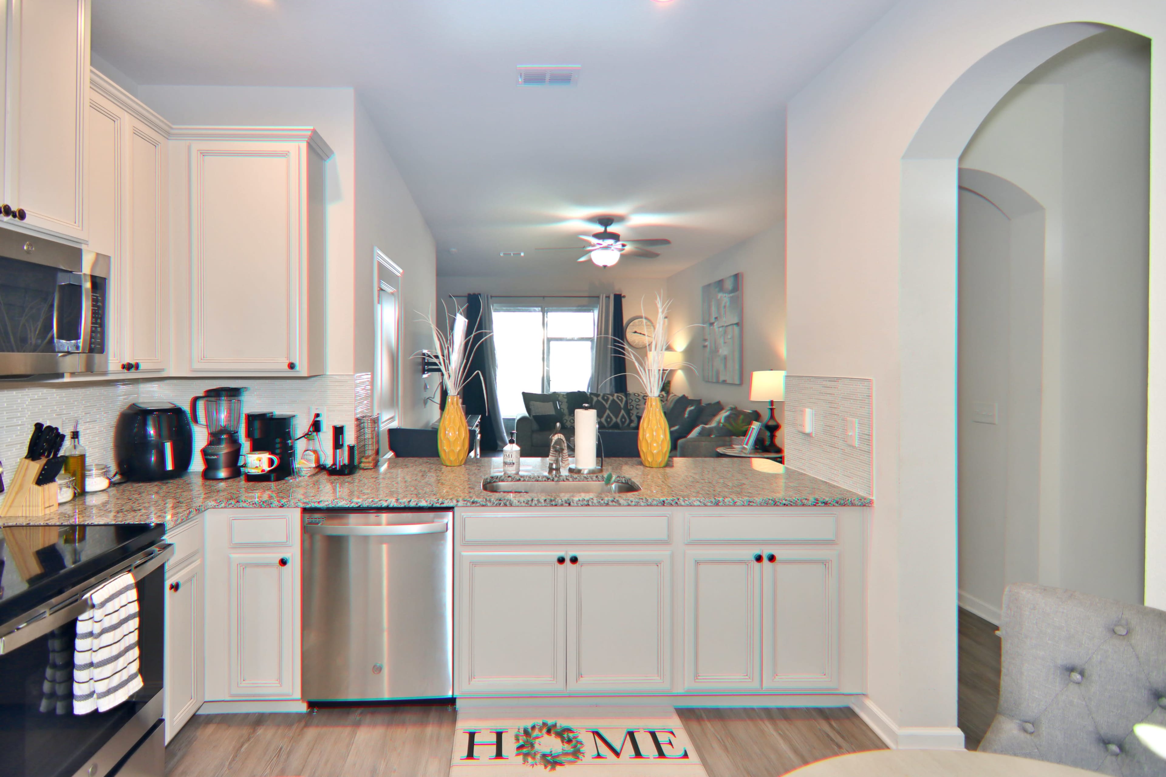 A modern kitchen featuring light-colored cabinets, a stainless steel dishwasher, and a view into a living area with a ceiling fan and dark furnishings.