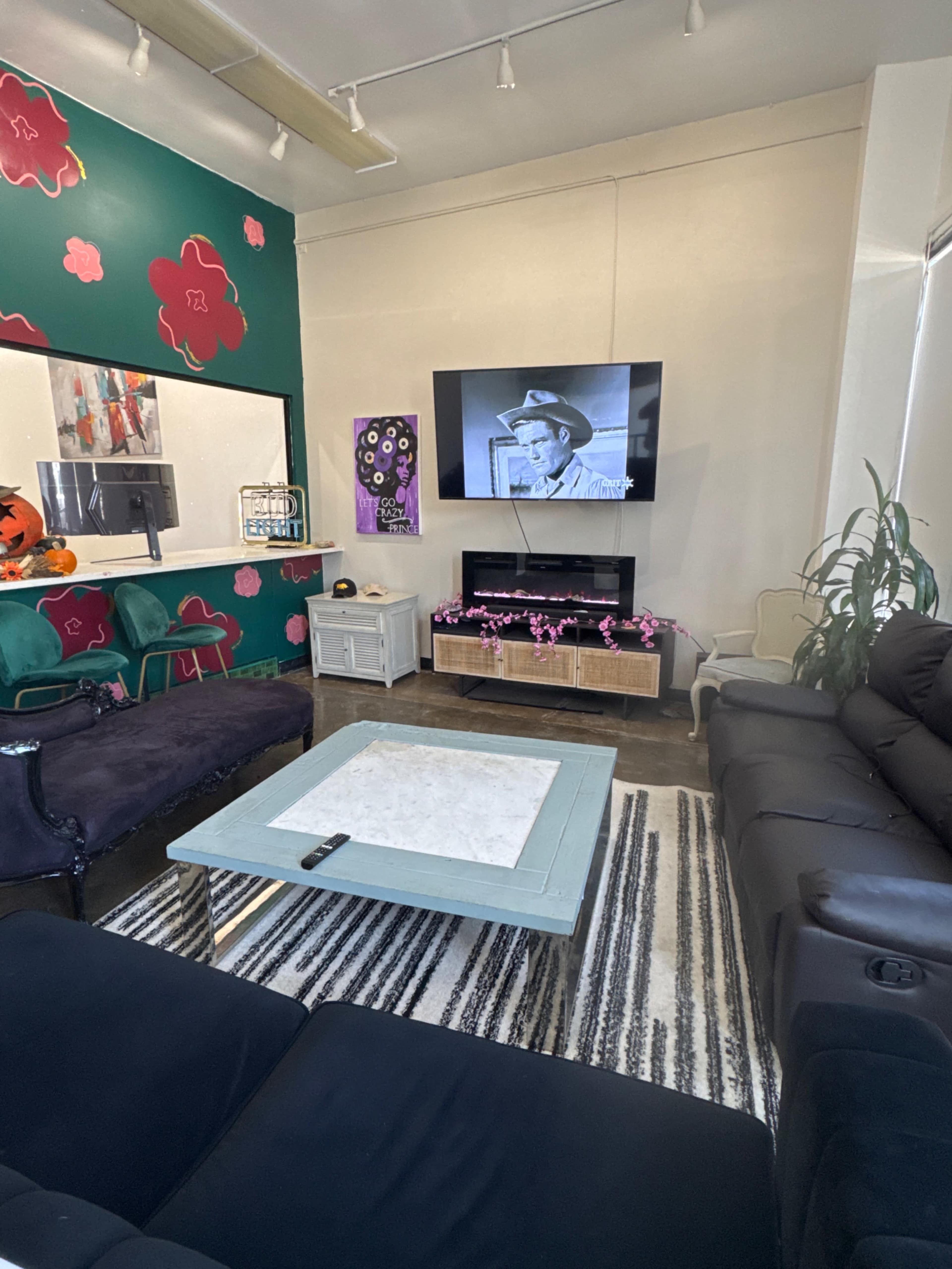 The image shows a cozy lounge area with two couches, a television mounted on the wall, and a coffee table on a striped rug, decorated with purple flowers and other accessories.