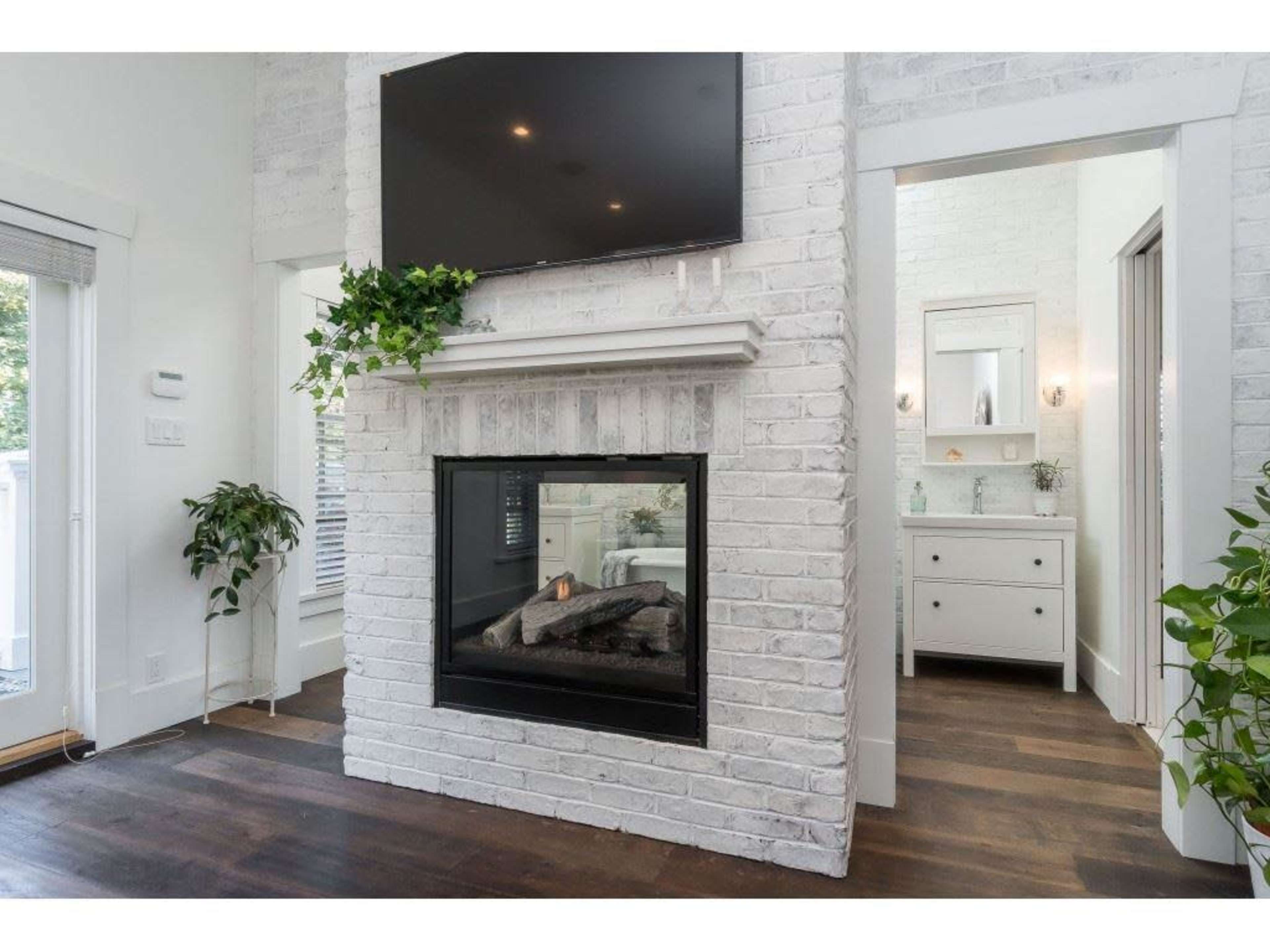 The image shows a modern living space with a white brick fireplace, a mounted television above it, and a view into a bathroom area with a vanity.
