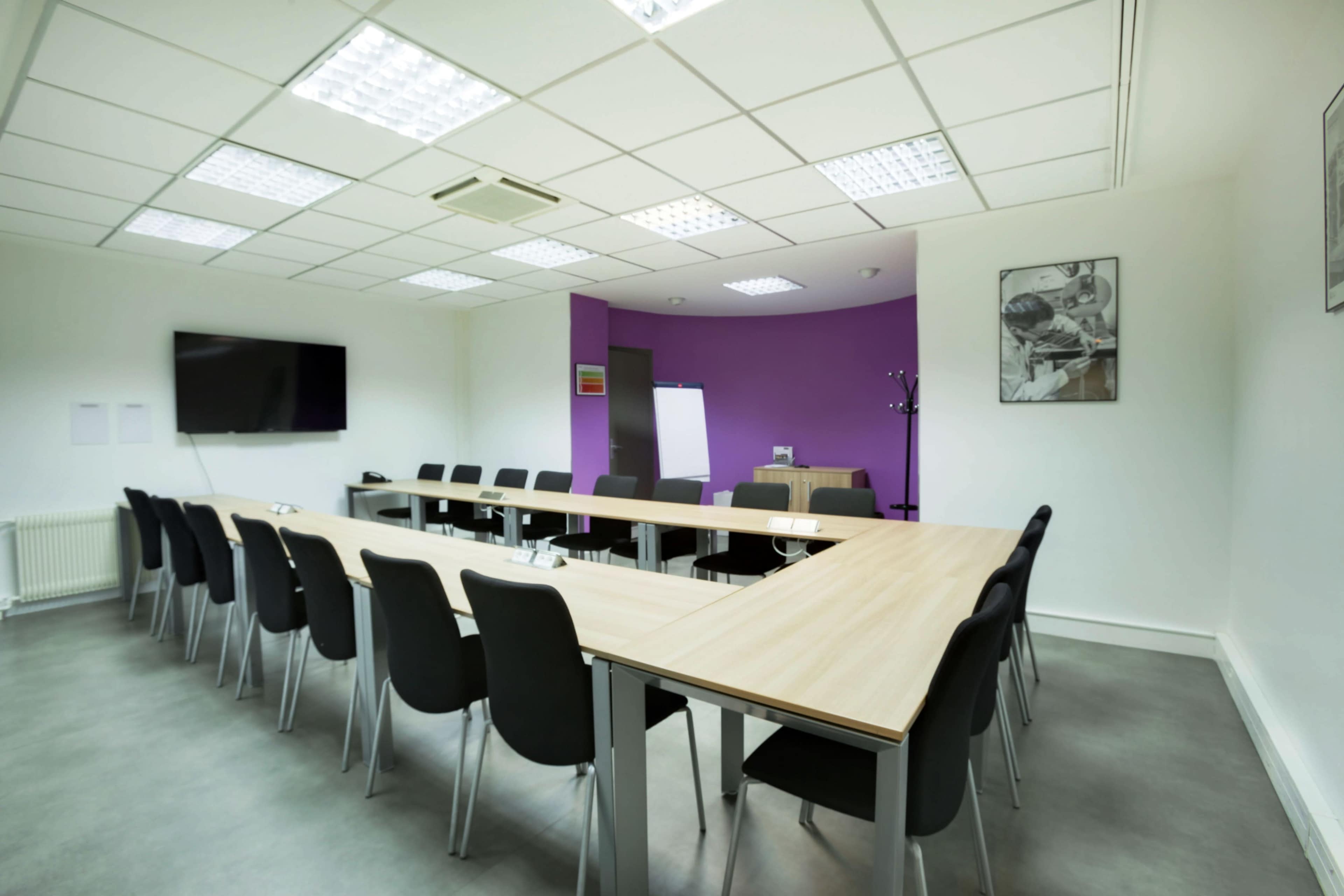A conference room is arranged with a long table and black chairs, featuring a large screen and a purple accent wall.
