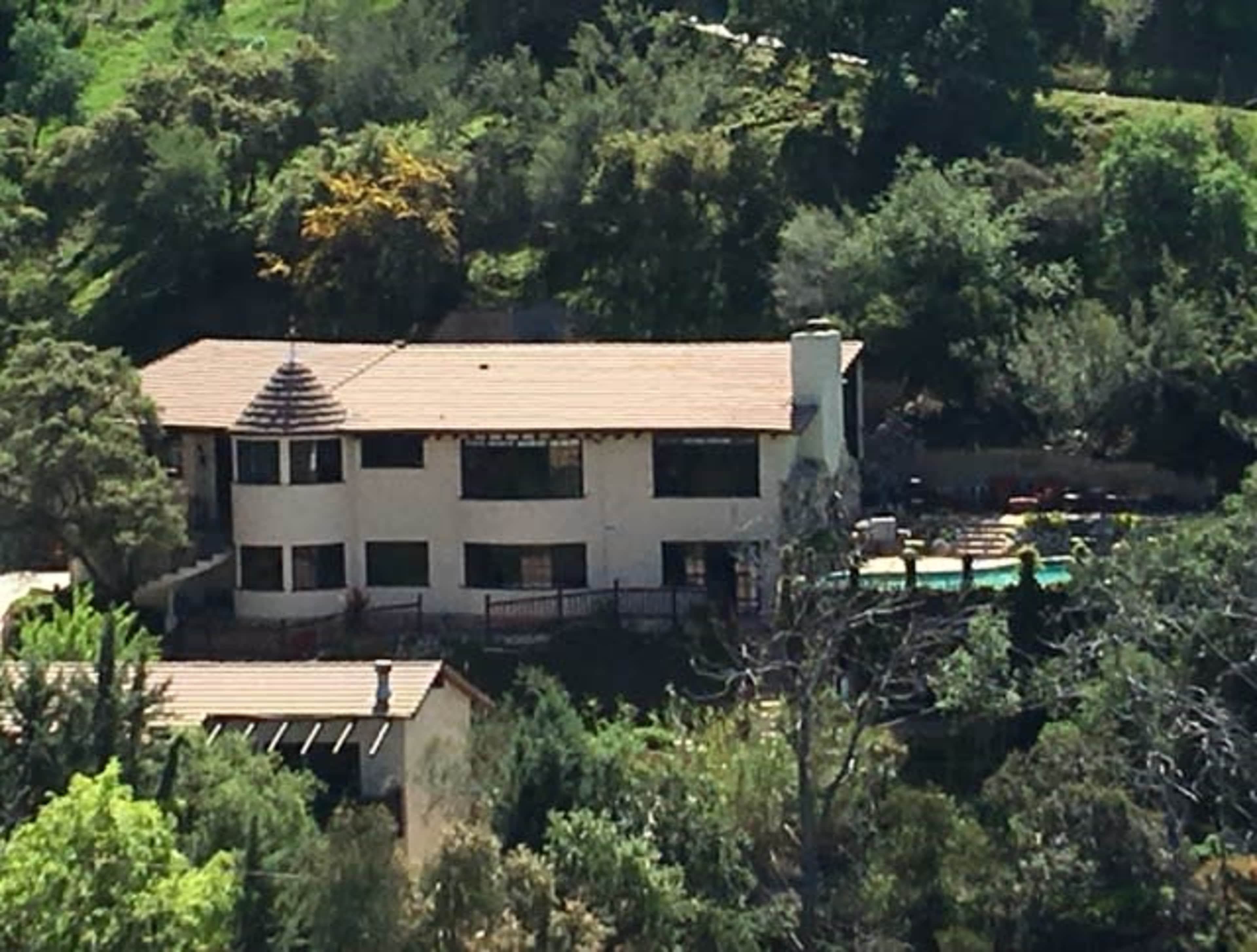 A large, two-story house with a tiled roof is situated amidst greenery, featuring a swimming pool and outdoor seating area.