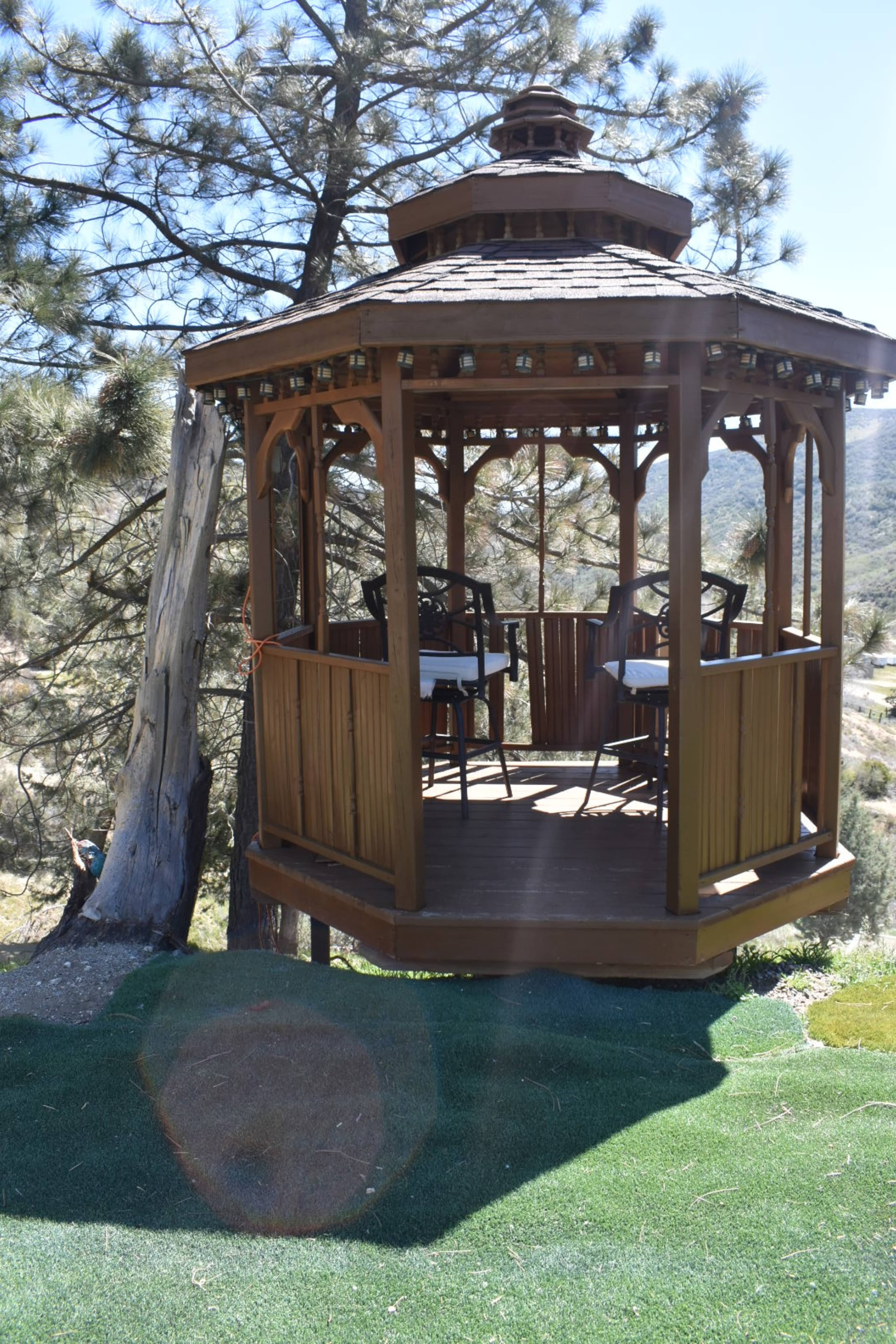 The image shows a wooden gazebo with a circular seating area set on a grassy slope, surrounded by trees and mountains in the background.
