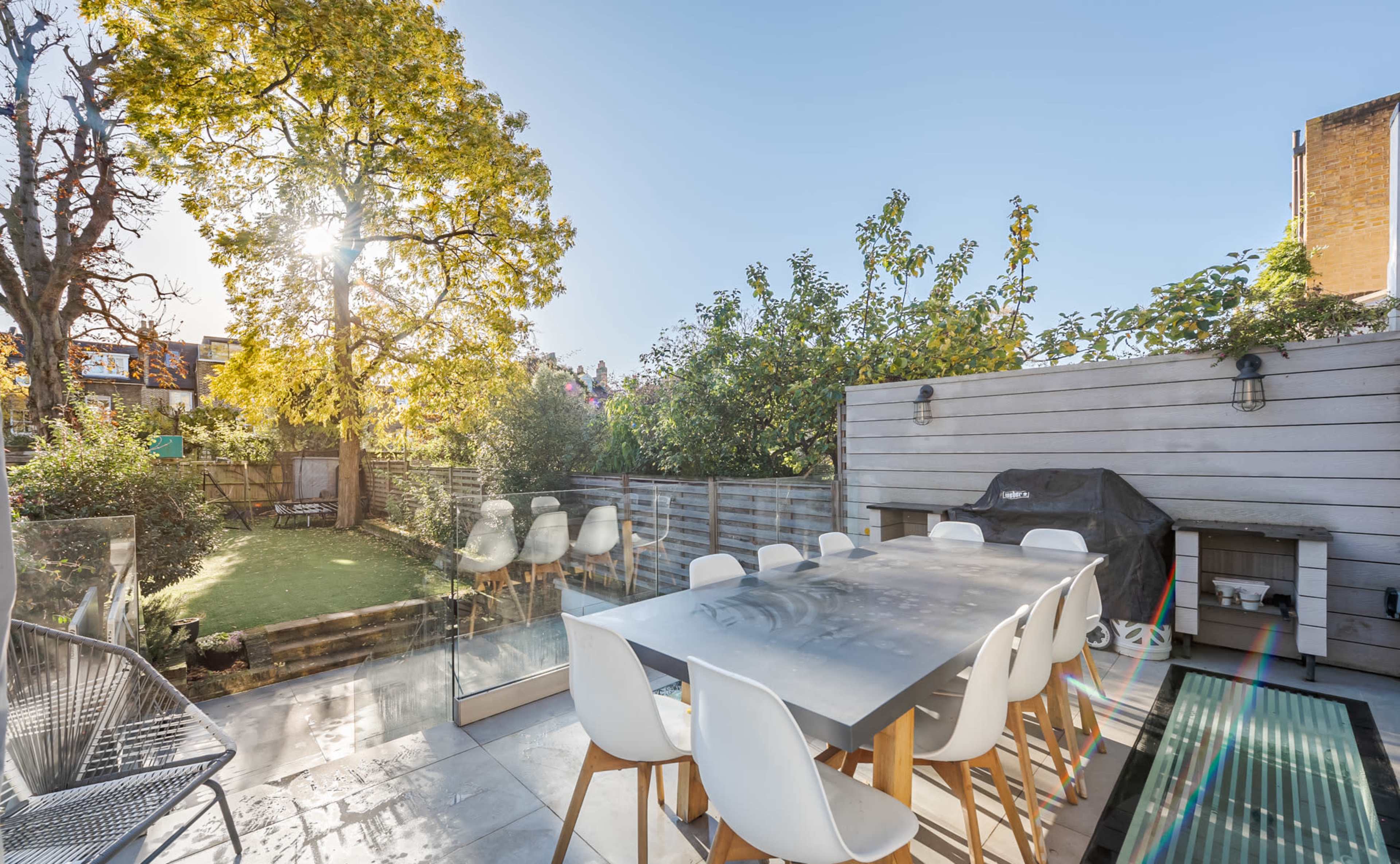 A modern outdoor dining area features a large table surrounded by white chairs, with a lush garden and towering trees in the background.