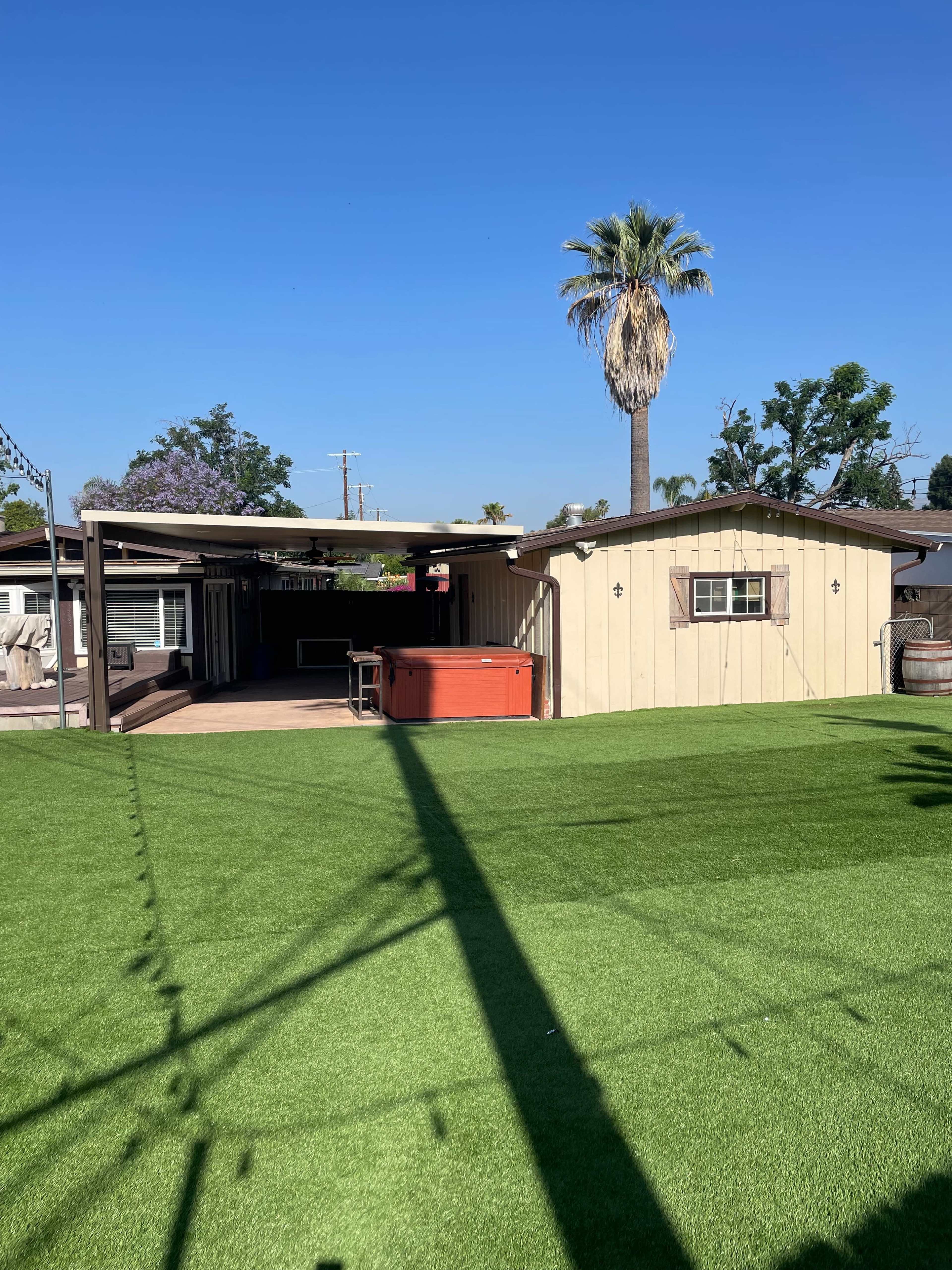 A backyard features a lawn of artificial turf, a palm tree, and a wooden structure with a hot tub.
