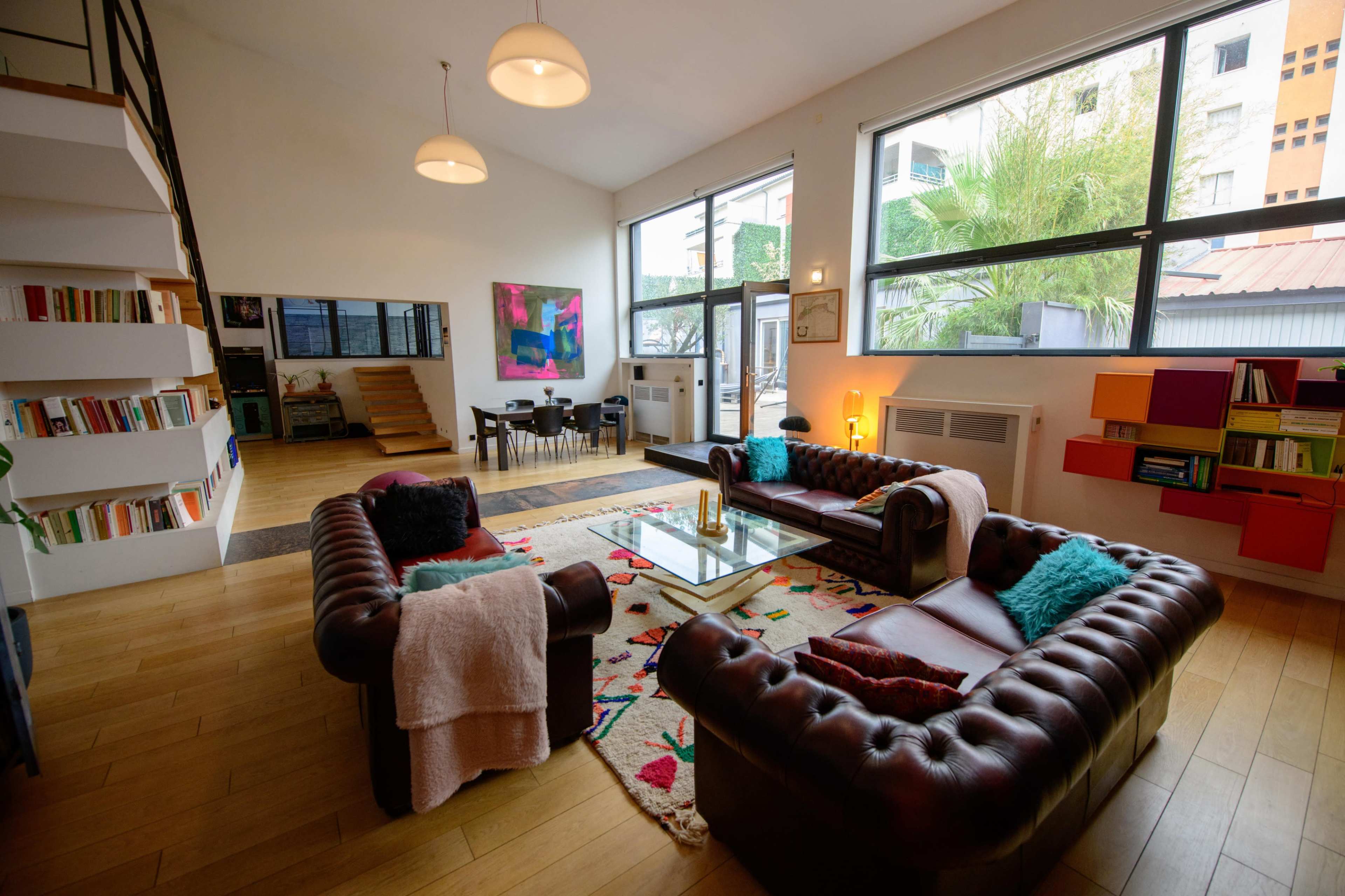 The image shows a modern living room with a leather sofa set, a coffee table, large windows, and a staircase leading to an upper level.