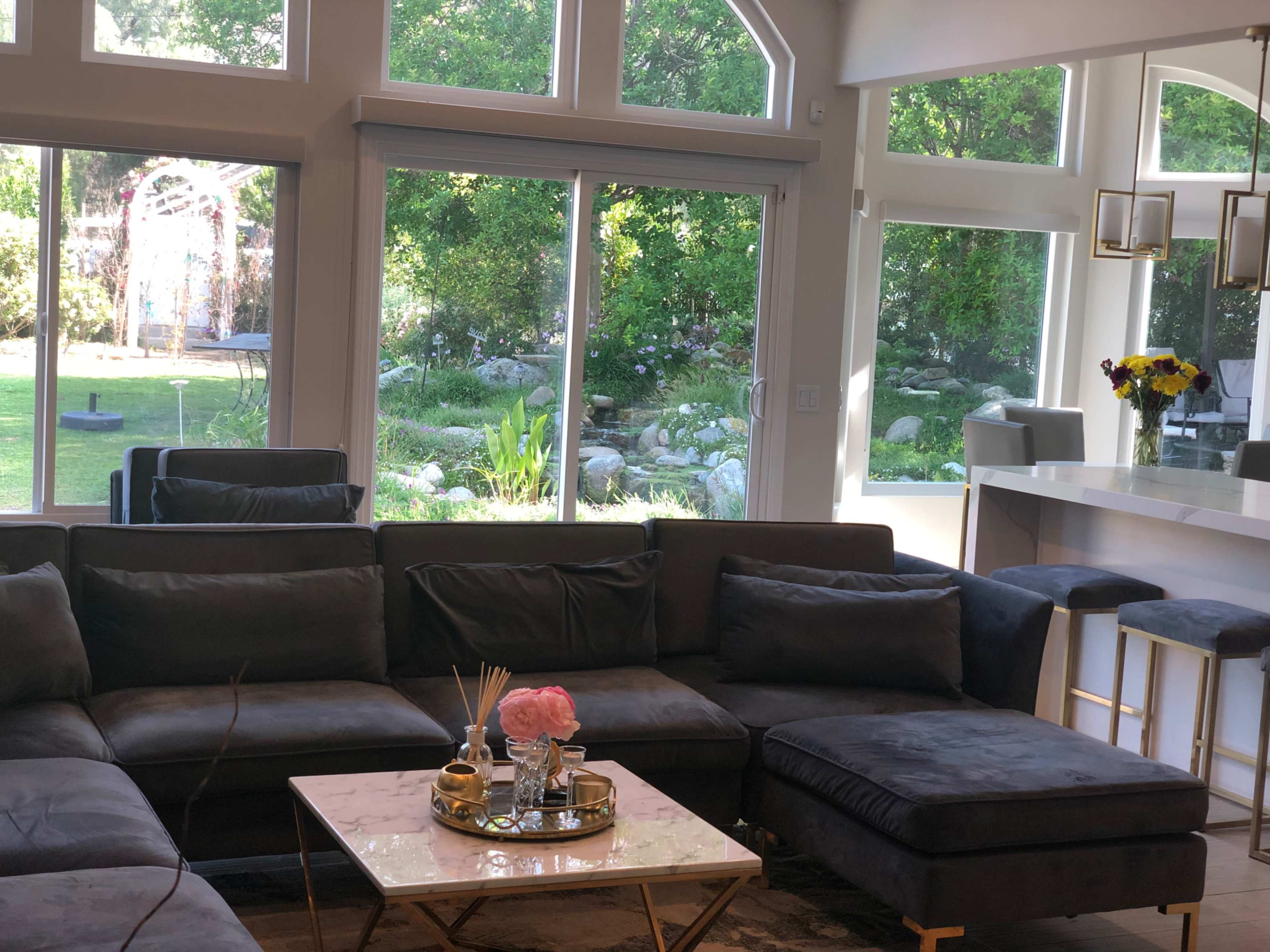 A modern living room with a dark sectional sofa, a coffee table, and large windows that overlook a garden with greenery and a decorative archway.