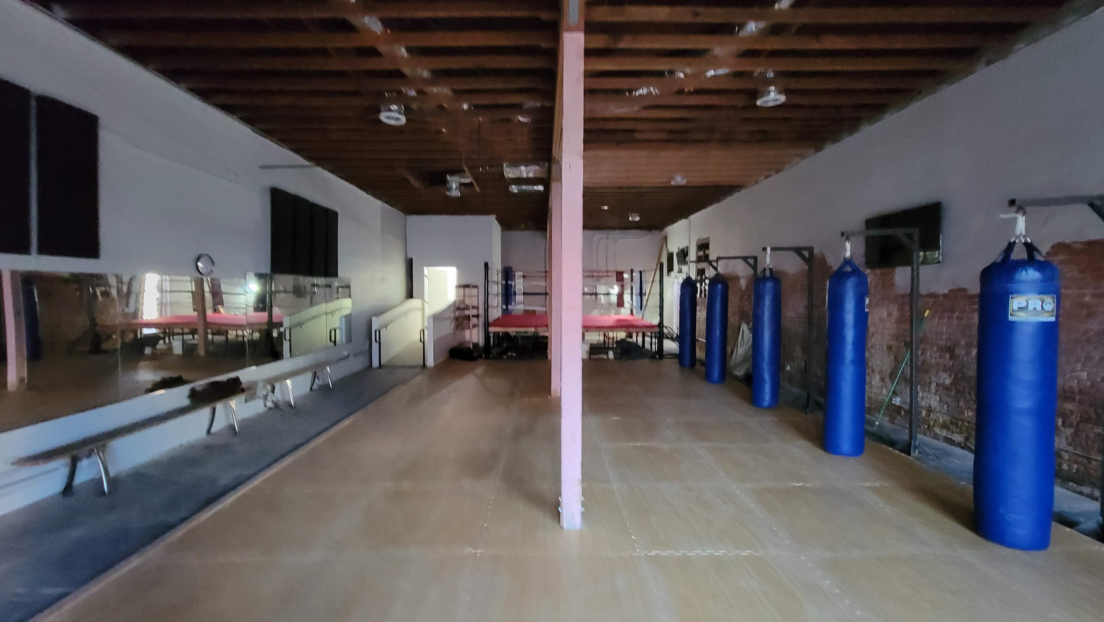 An empty boxing gym with a central wooden floor, blue punching bags hanging on the walls, and a boxing ring in the background.