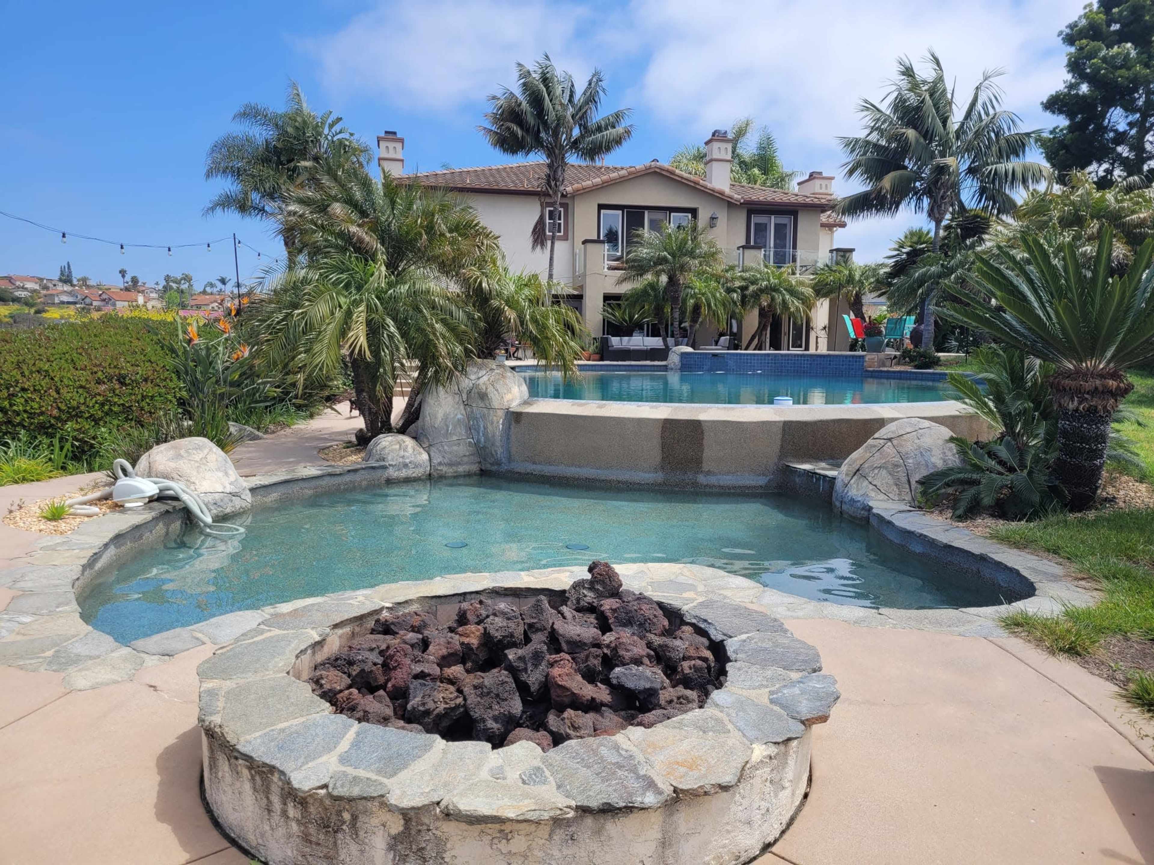 The image shows a luxurious backyard with a swimming pool and a fire pit surrounded by tropical plants and a large house in the background.