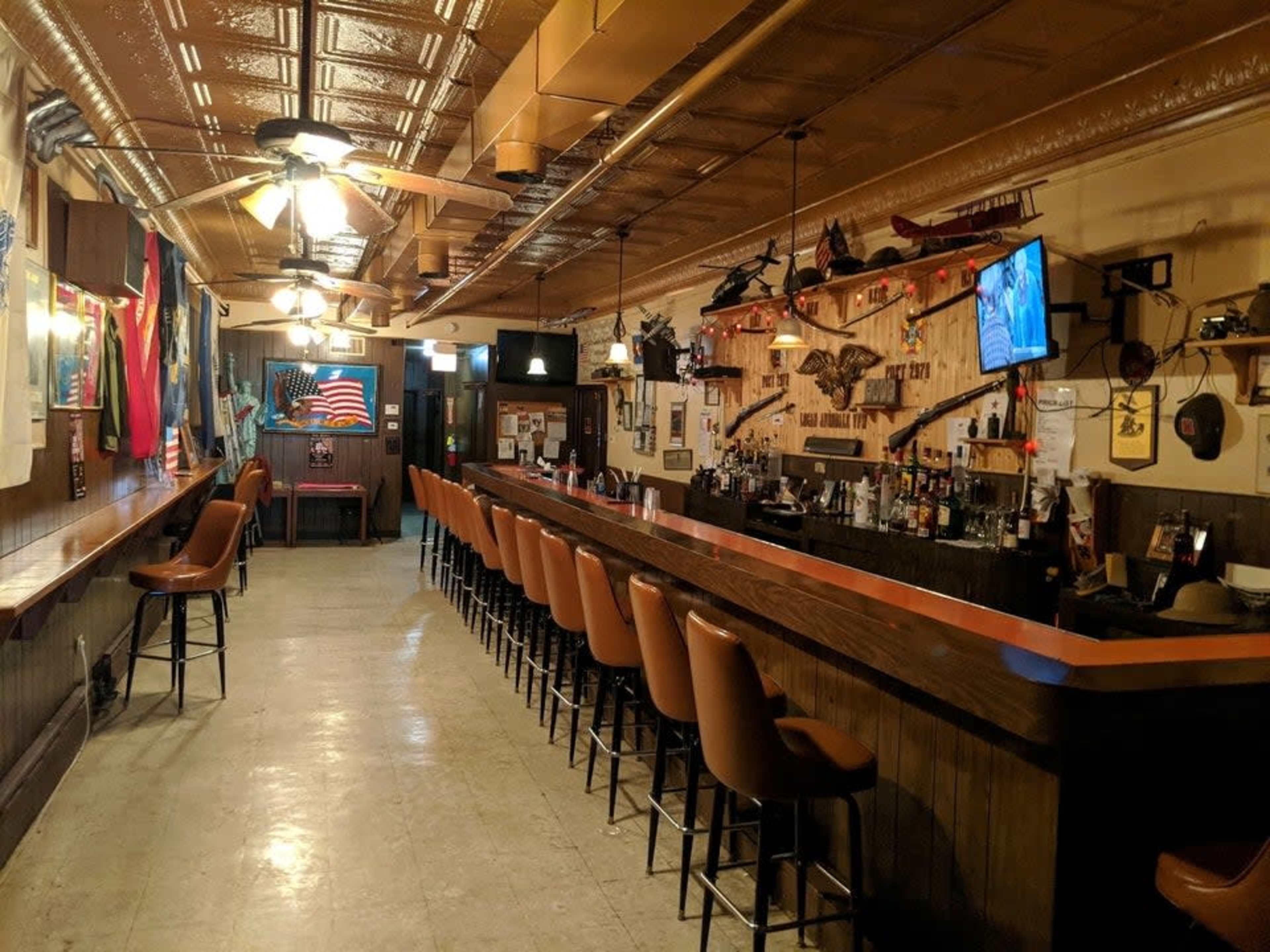 The interior of a bar with a long wooden counter, several high-backed seats, and various decorations, including flags and wall-mounted memorabilia.