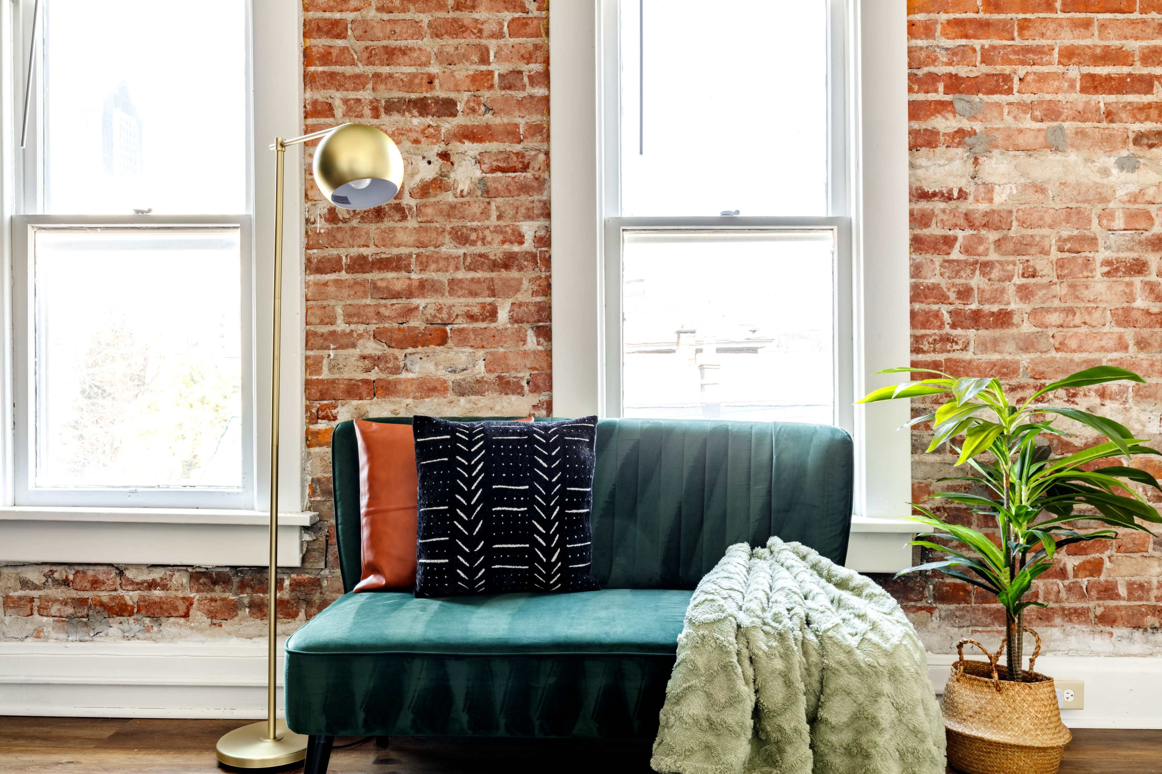 A green sofa with patterned cushions is positioned against a brick wall, accompanied by a floor lamp and a potted plant.