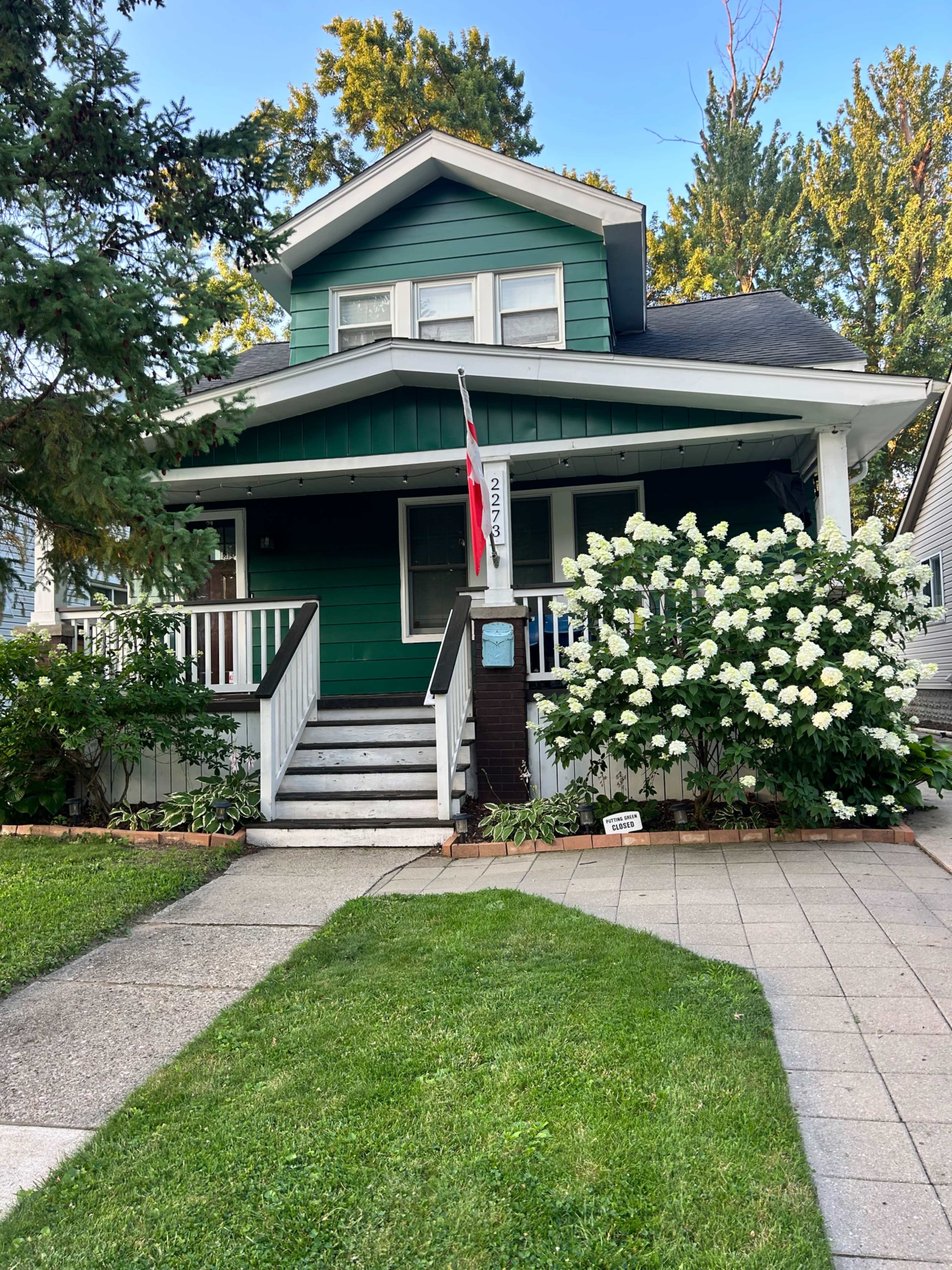 A green two-story house with a front porch and white trim is surrounded by lush greenery and blooming white flowers.
