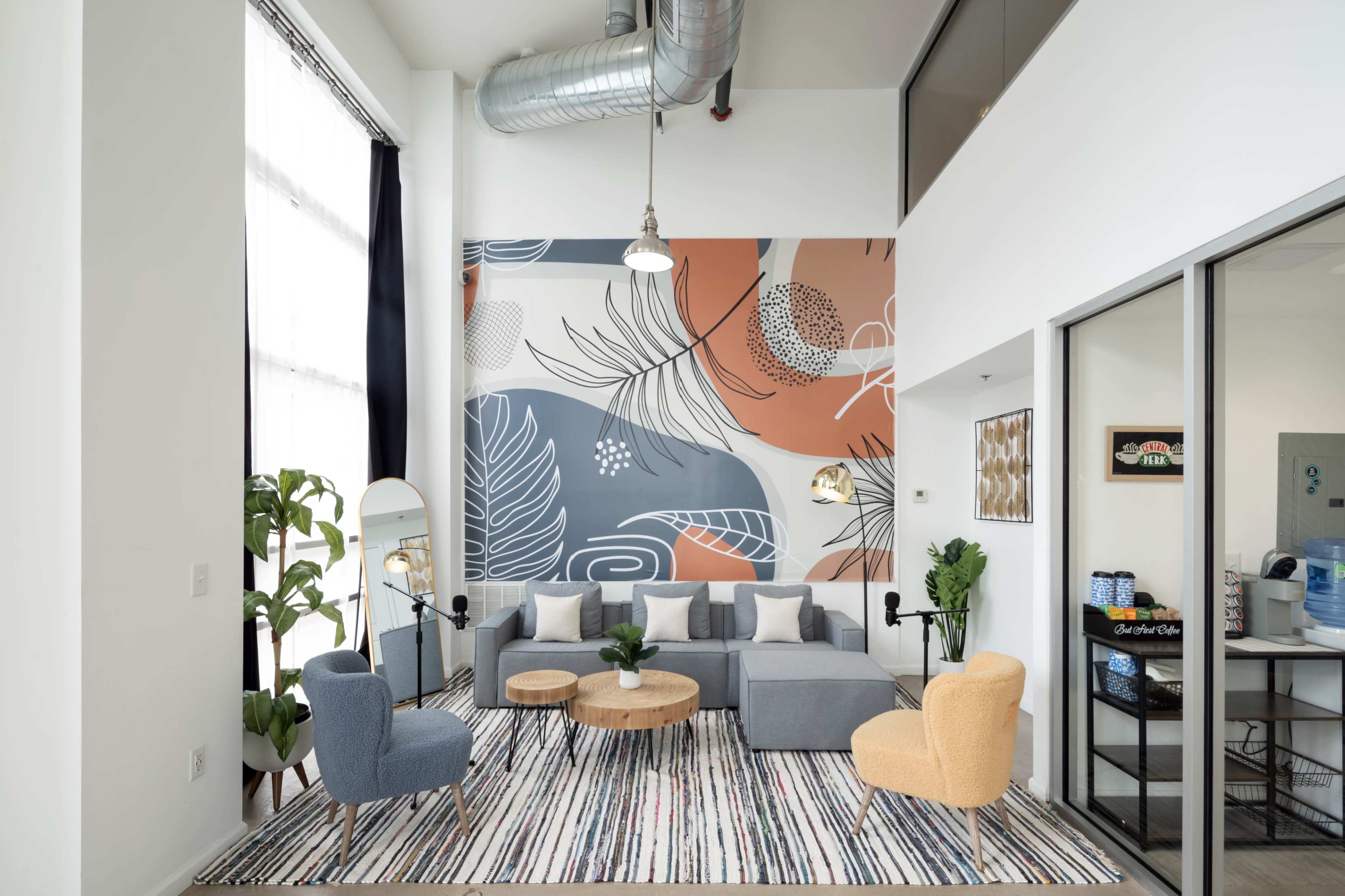 A modern living room decorated with a patterned accent wall, a gray sectional sofa, two colorful armchairs, a round wooden coffee table, and various indoor plants.