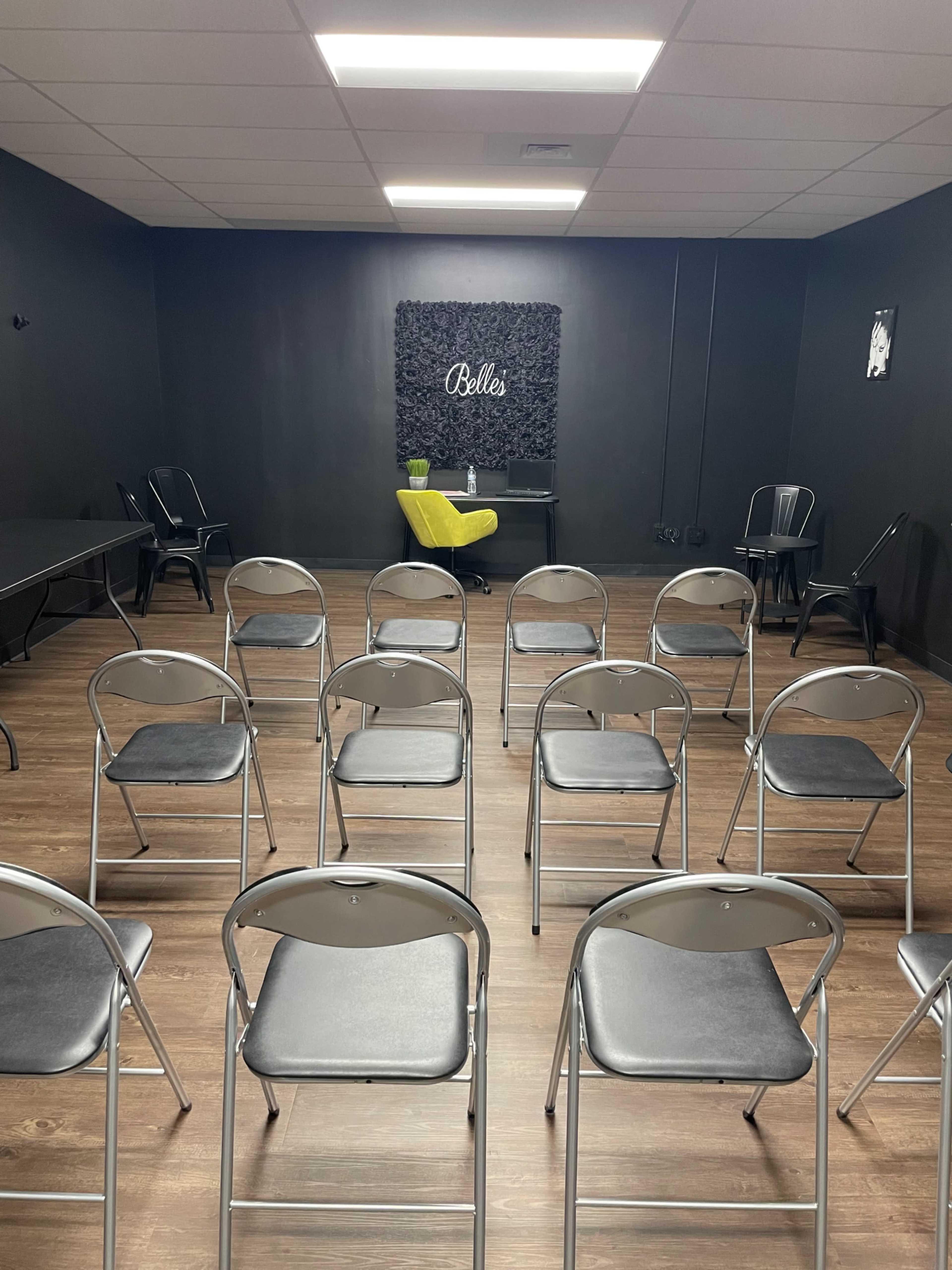A room set up with rows of empty metal folding chairs facing a small, yellow chair and a table against a dark wall adorned with a decorative sign that reads "Belles."