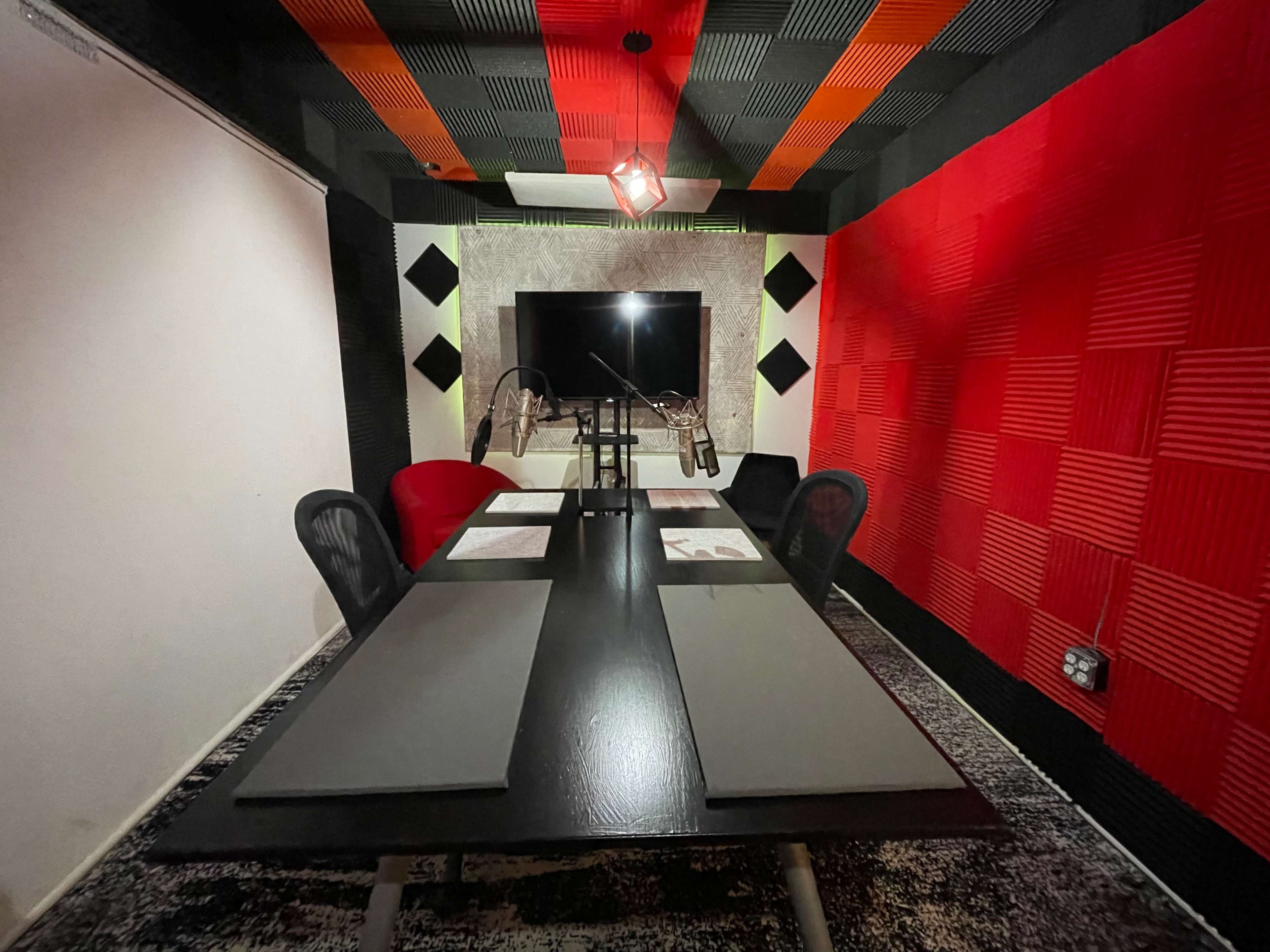 A soundproof recording studio features a long black table with chairs, red and black acoustic foam on the walls, and a large screen at the far end.