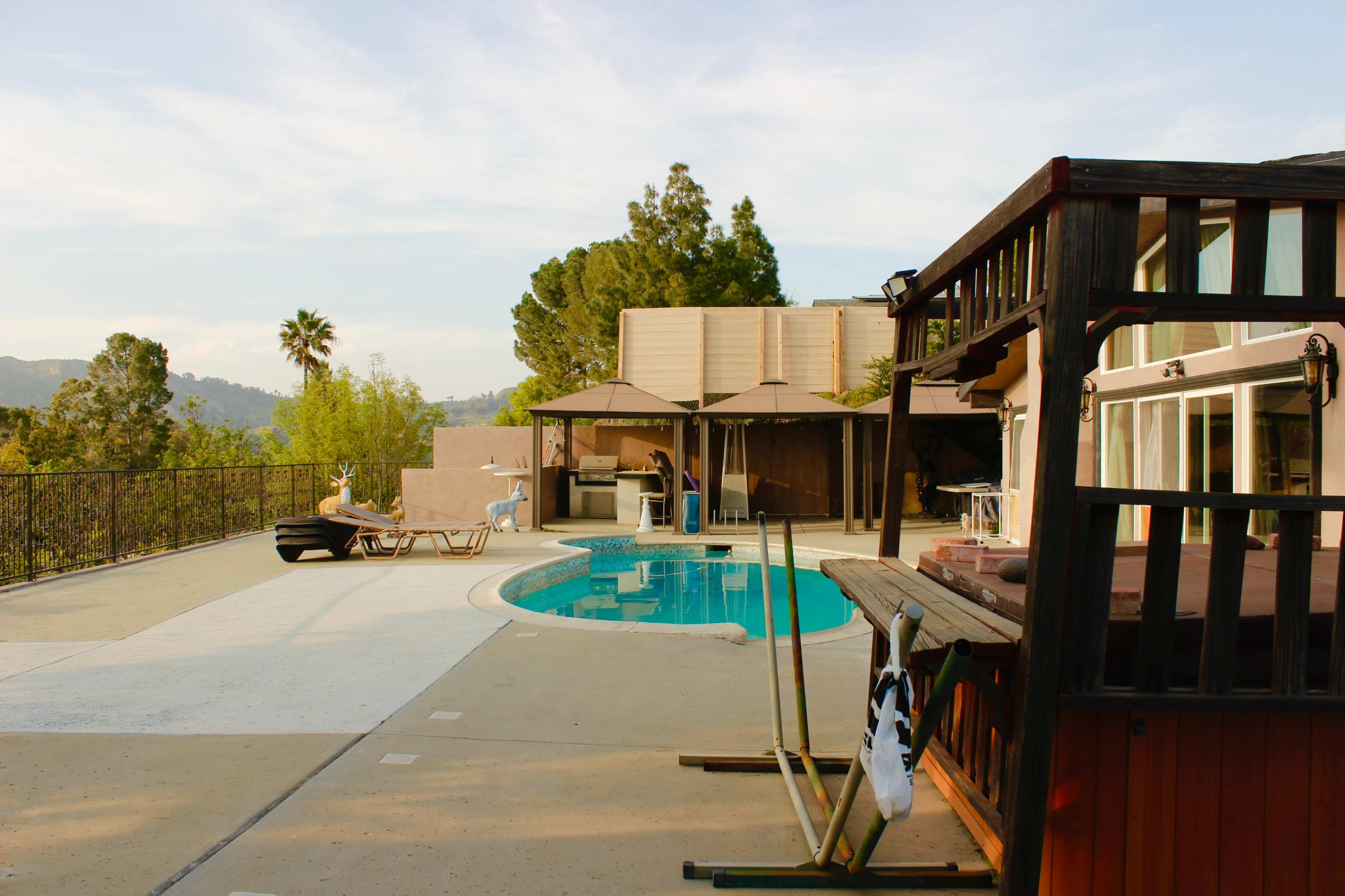 A spacious outdoor area featuring a swimming pool, lounge chairs, and a wooden structure, set against a backdrop of trees and hills.