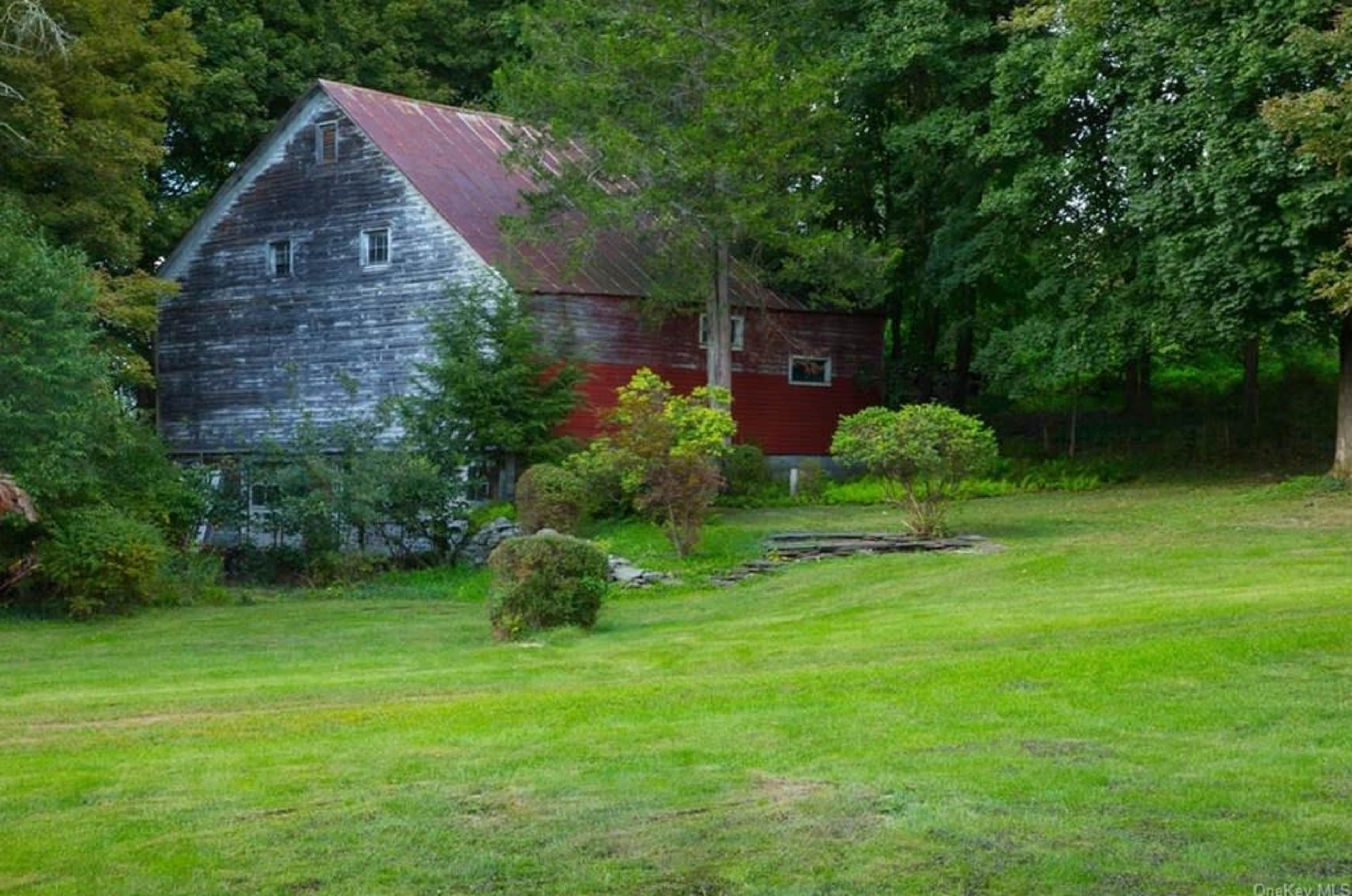 A weathered barn with a red section stands in a grassy area surrounded by trees and foliage.