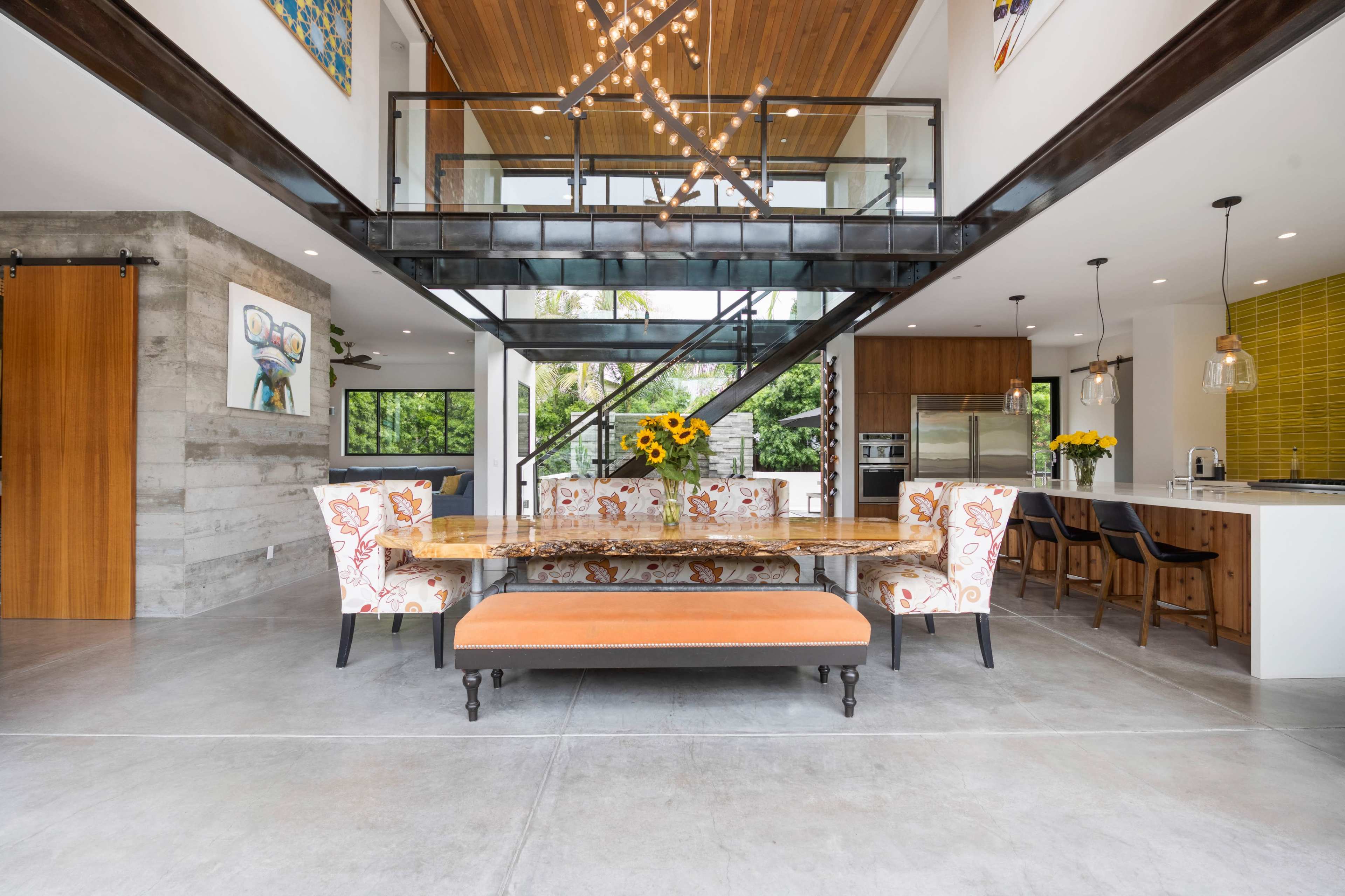 A modern, open-concept dining area featuring a large wooden table surrounded by floral-patterned chairs and a bench, under a ceiling with wooden beams and an artistic light fixture.