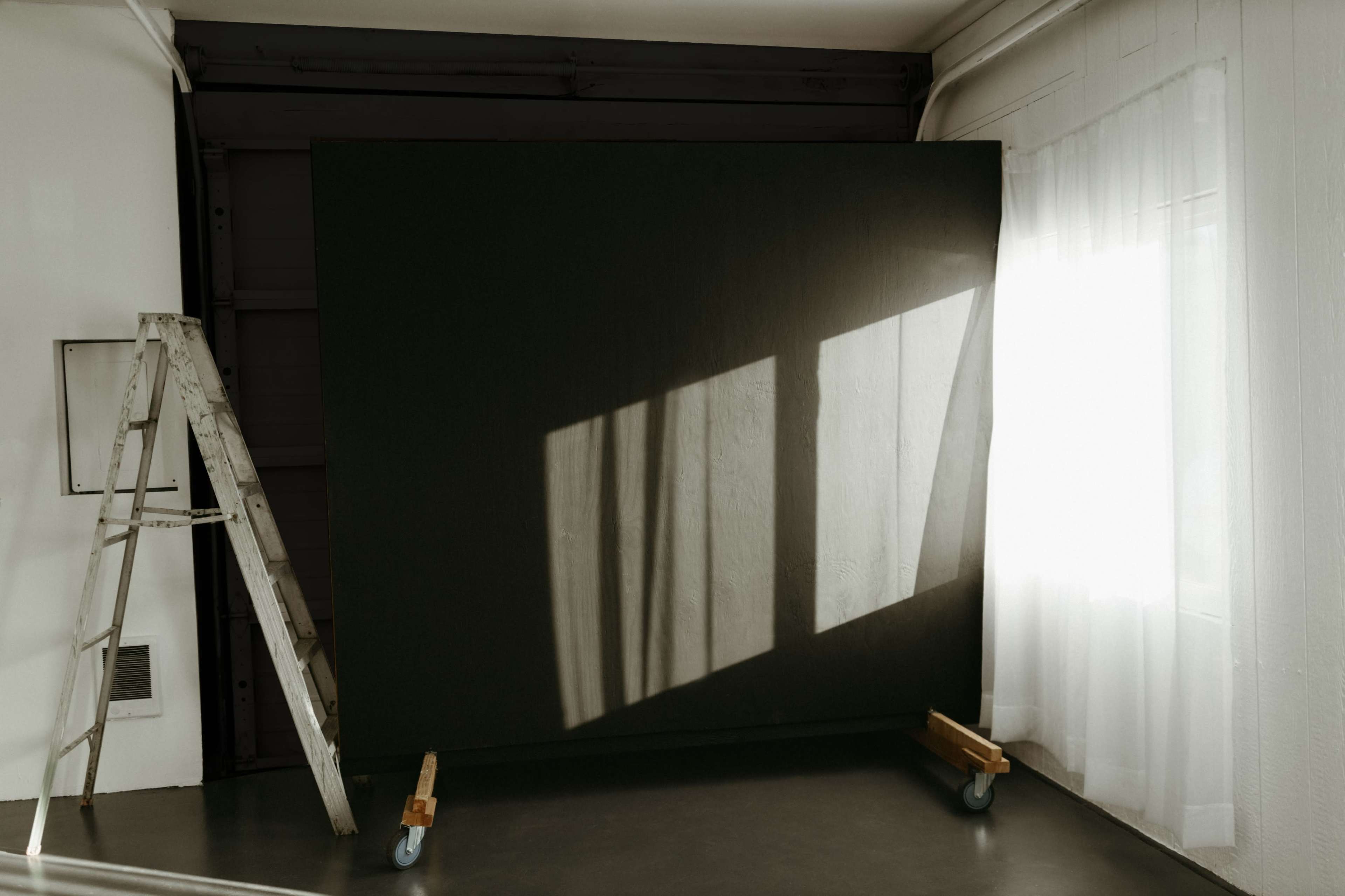 A large dark backdrop is positioned in a room with a ladder nearby, while sunlight casts shadows through a window onto the floor.