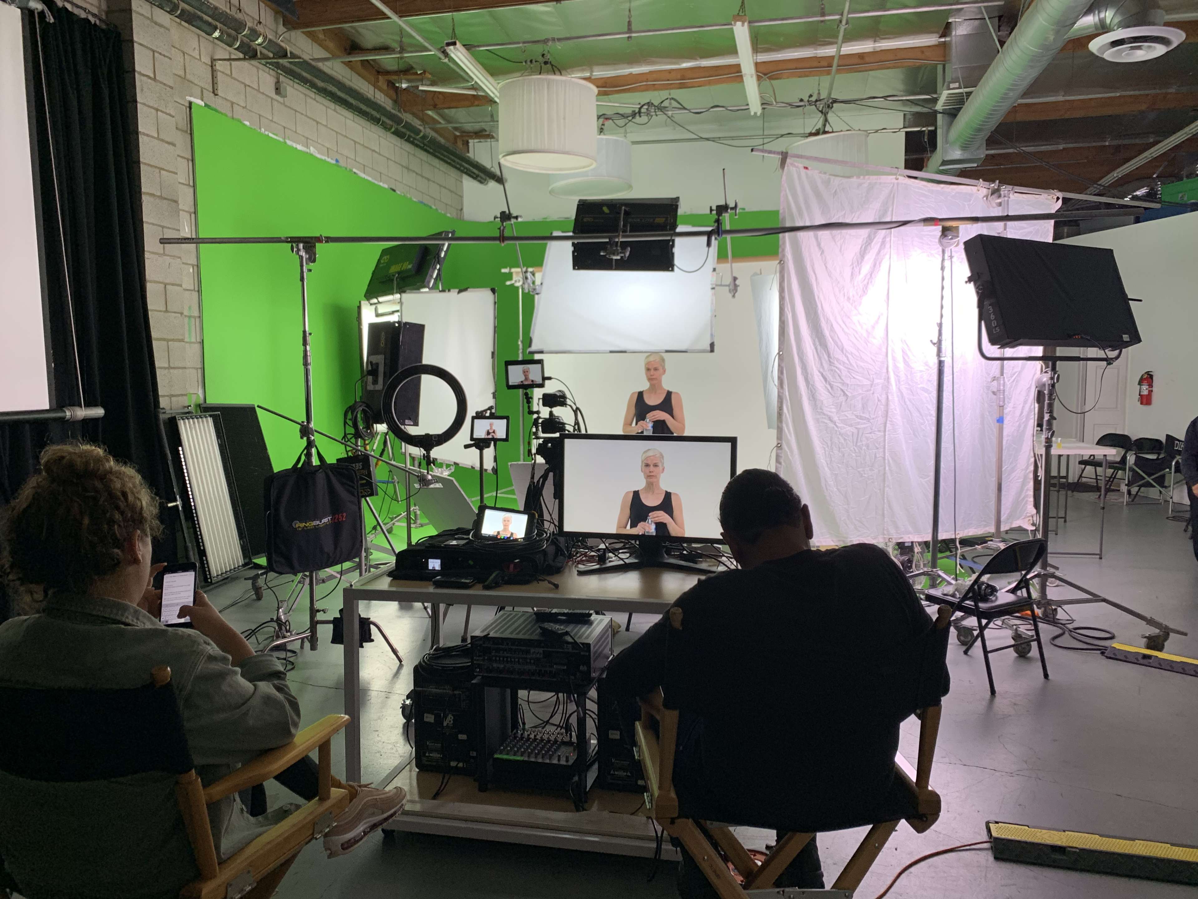 A film production setup featuring multiple cameras, lighting equipment, and a green screen with two people seated at a desk monitoring the scene.