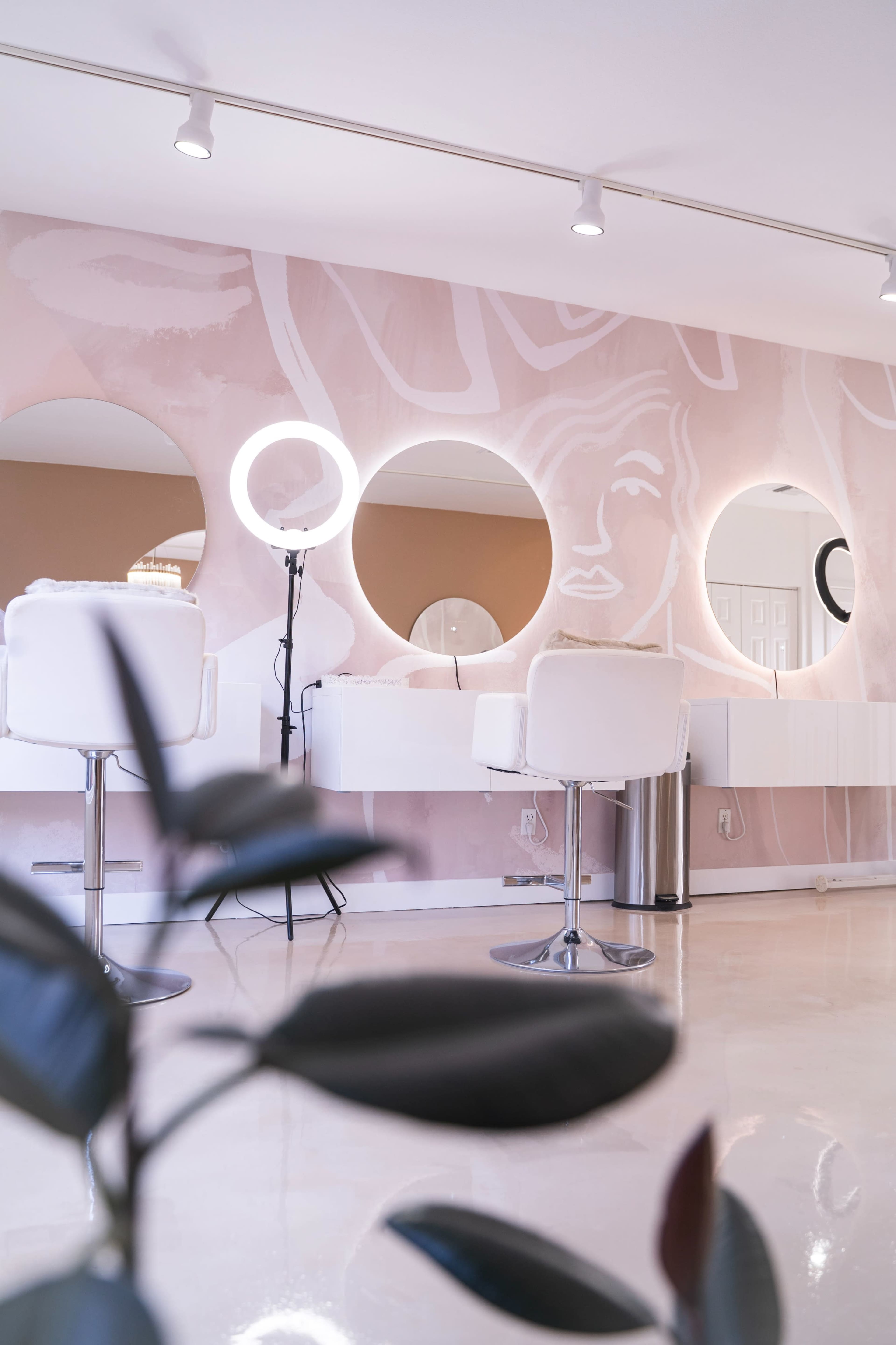 A modern hair salon interior with circular mirrors, sleek white chairs, and a minimalistic design featuring a pink mural on the wall.
