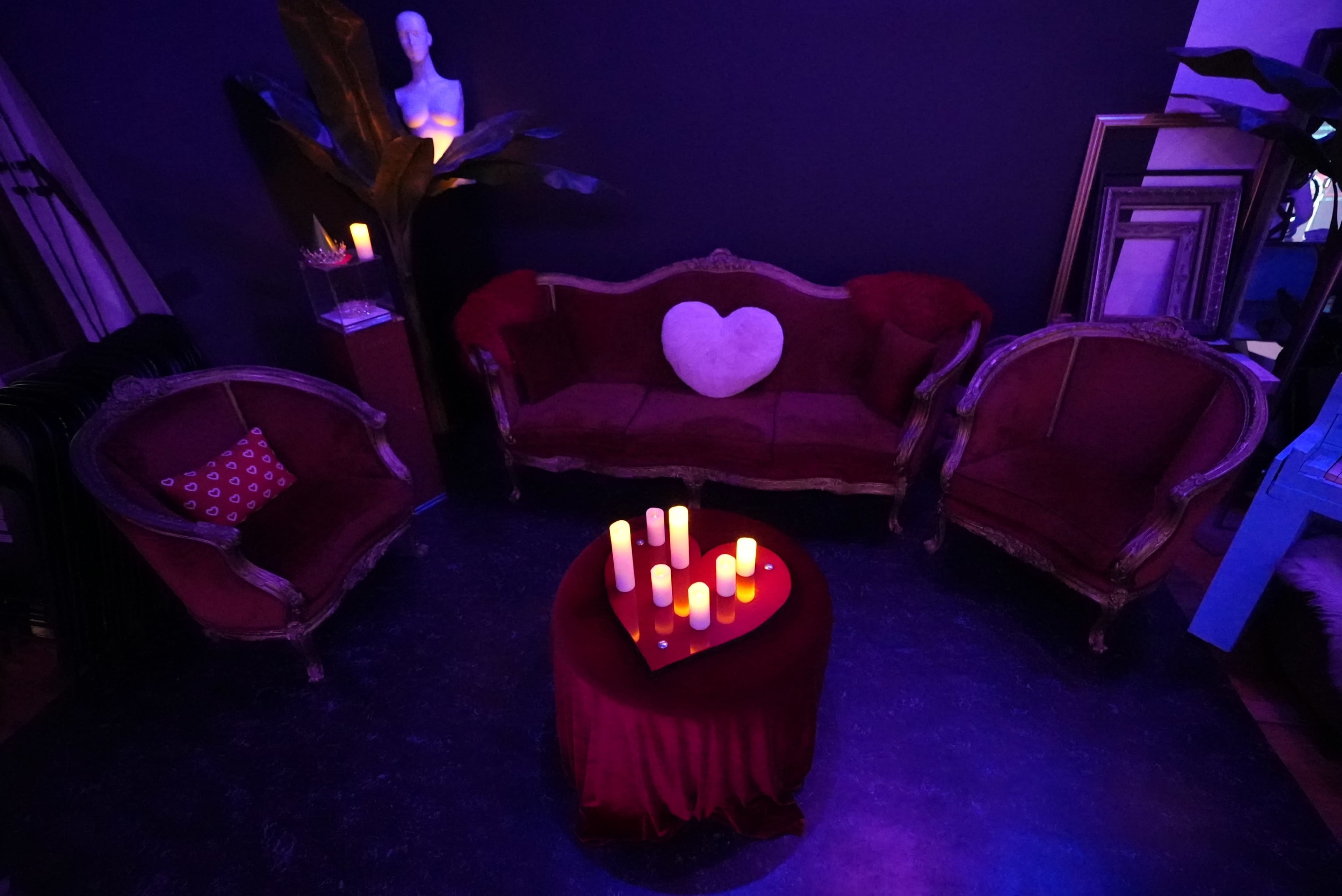 The image shows a dimly lit seating area featuring a red velvet sofa and armchairs arranged around a circular table with candles, all against a dark background.