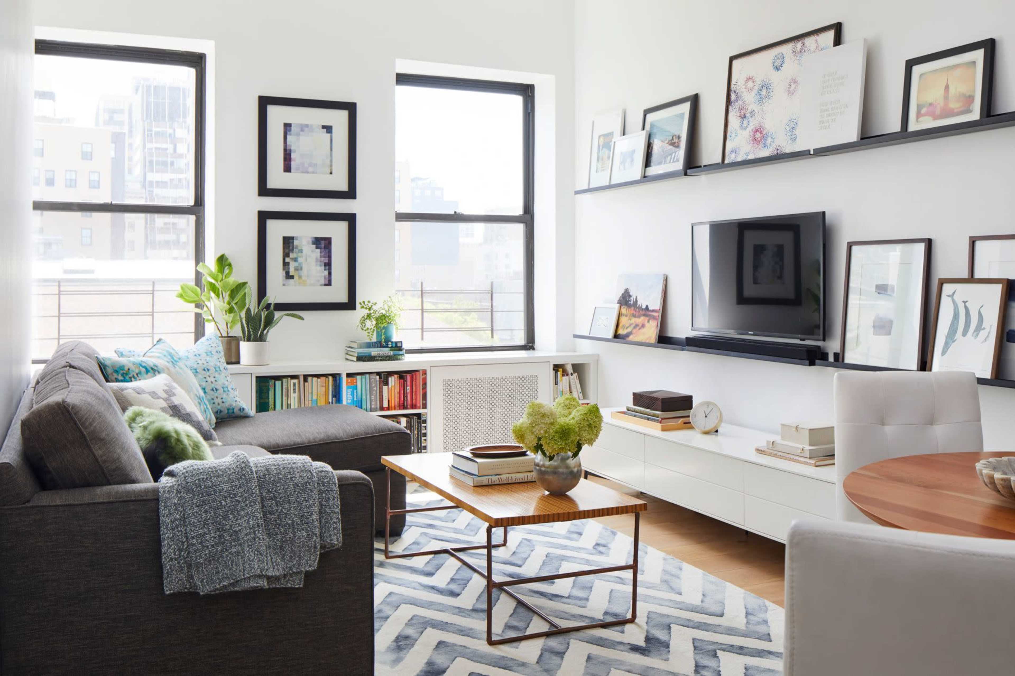 The living room features a modern design with a gray sofa, a wooden coffee table, wall shelves displaying framed art, and large windows allowing natural light to flood the space.