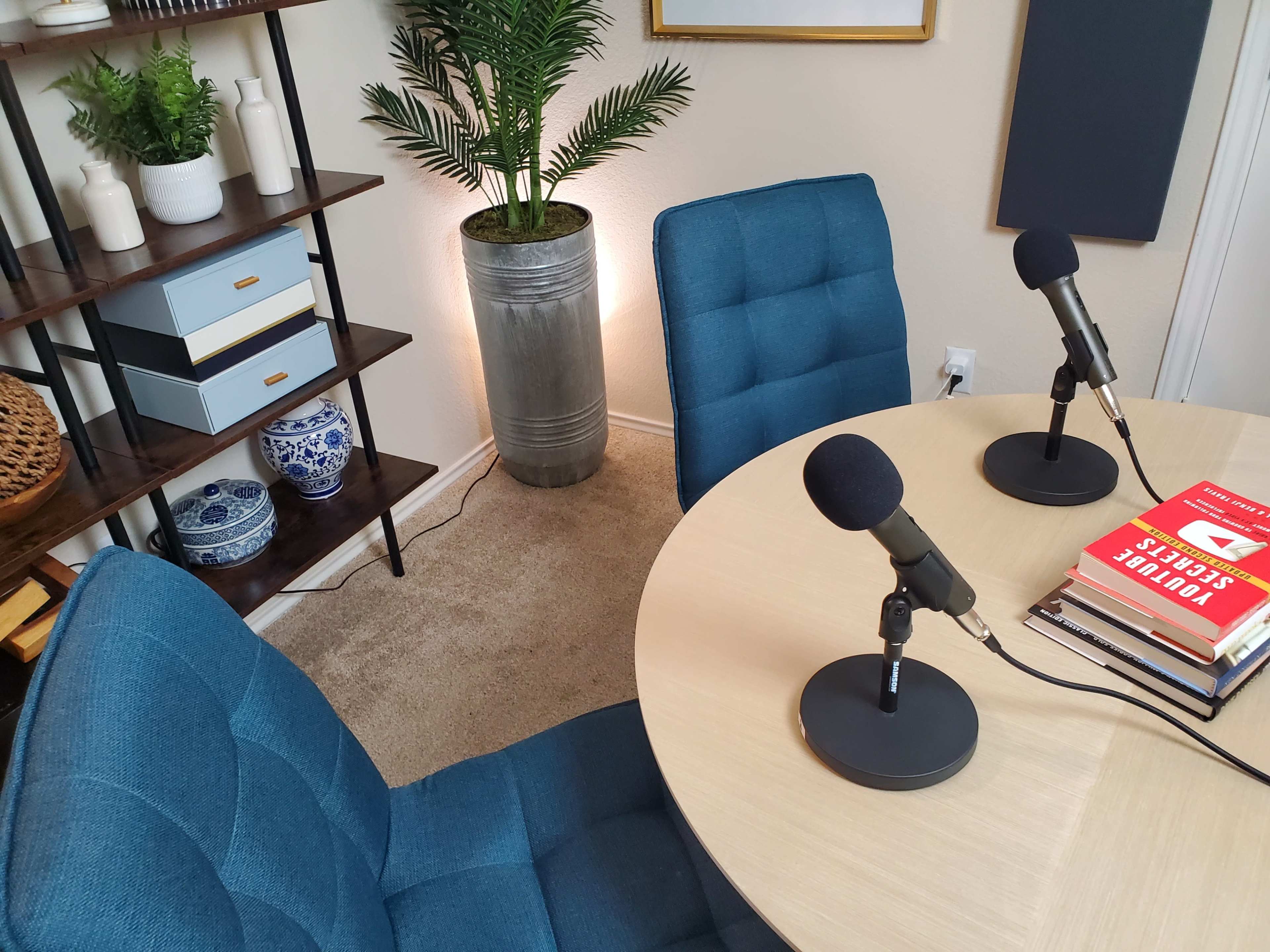 A podcast recording setup featuring two blue chairs, a round wooden table with a stack of books, and two microphones, surrounded by decorative plants and shelving.