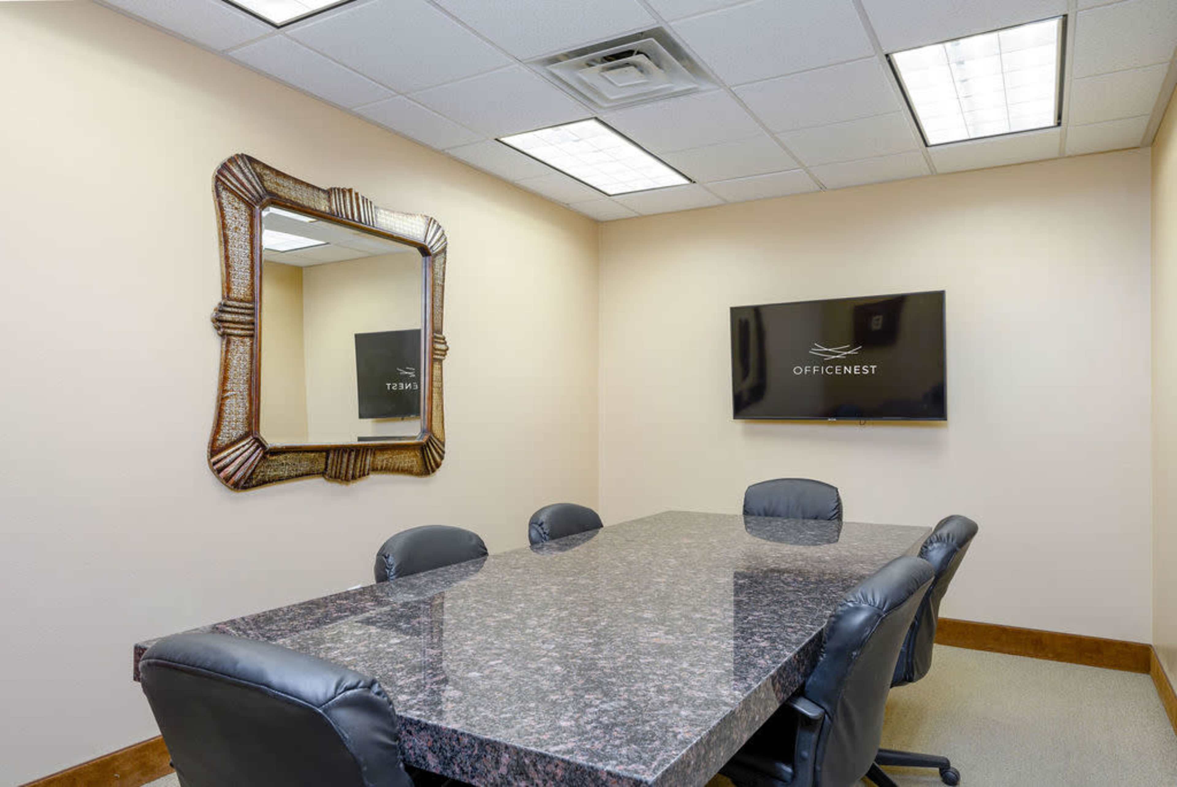 The image shows a small meeting room featuring a rectangular table surrounded by black leather chairs, a decorative mirror on the wall, and a wall-mounted TV displaying the logo "OFFICENEST."