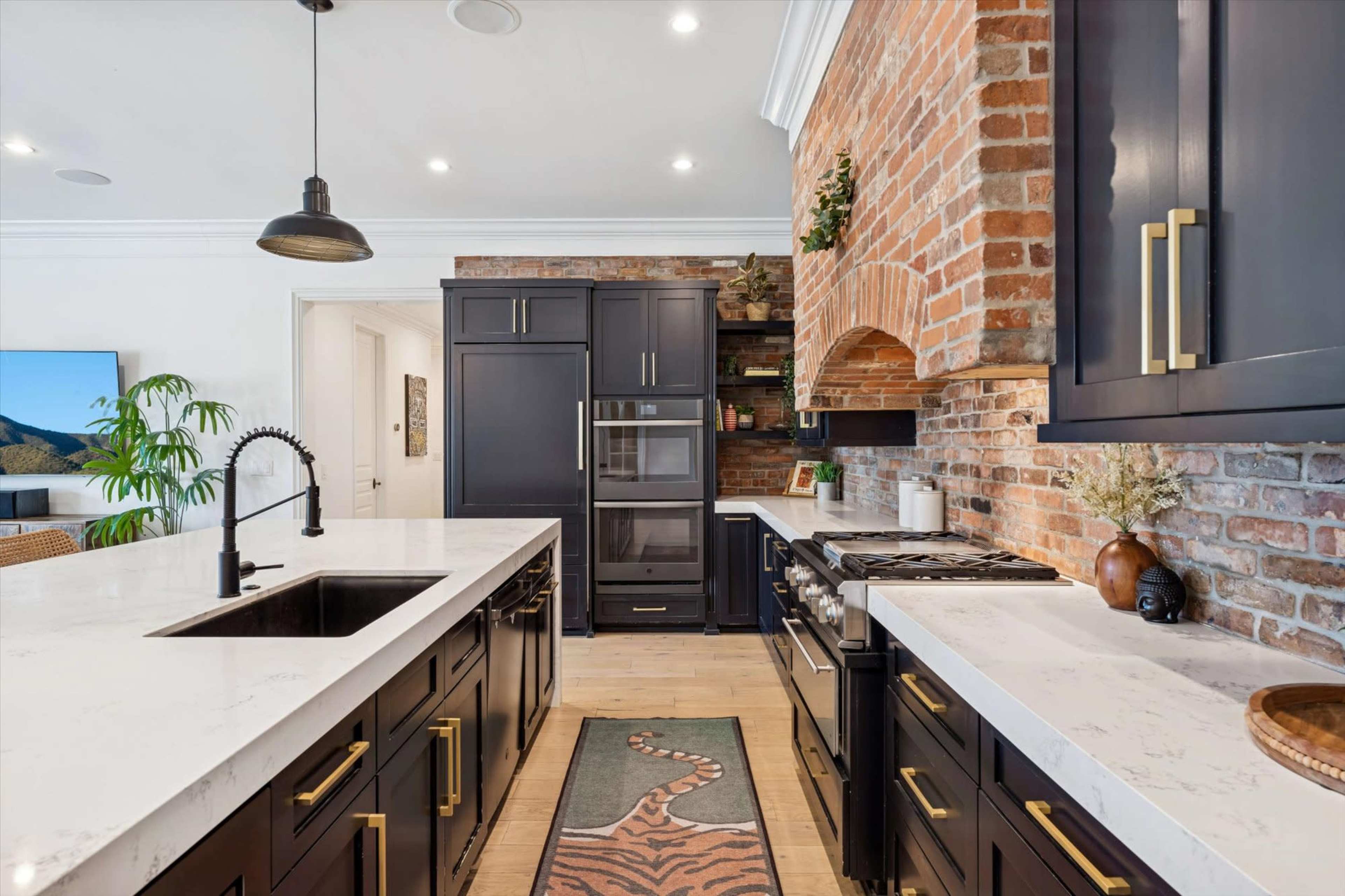 The image shows a modern kitchen featuring dark cabinetry, a brick accent wall, stainless steel appliances, and an island with white countertops.