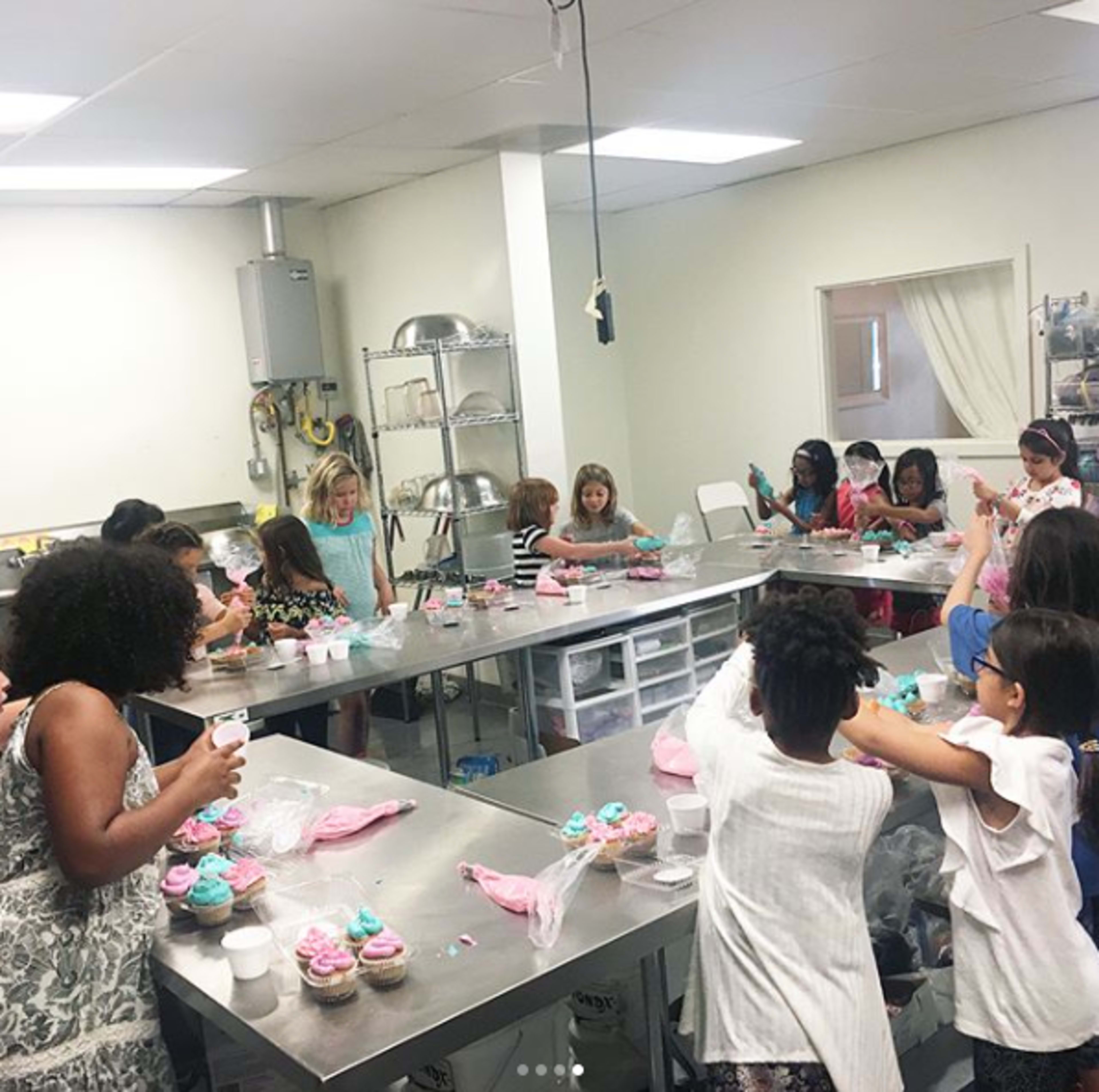 A group of children and adults is decorating cupcakes in a commercial kitchen.