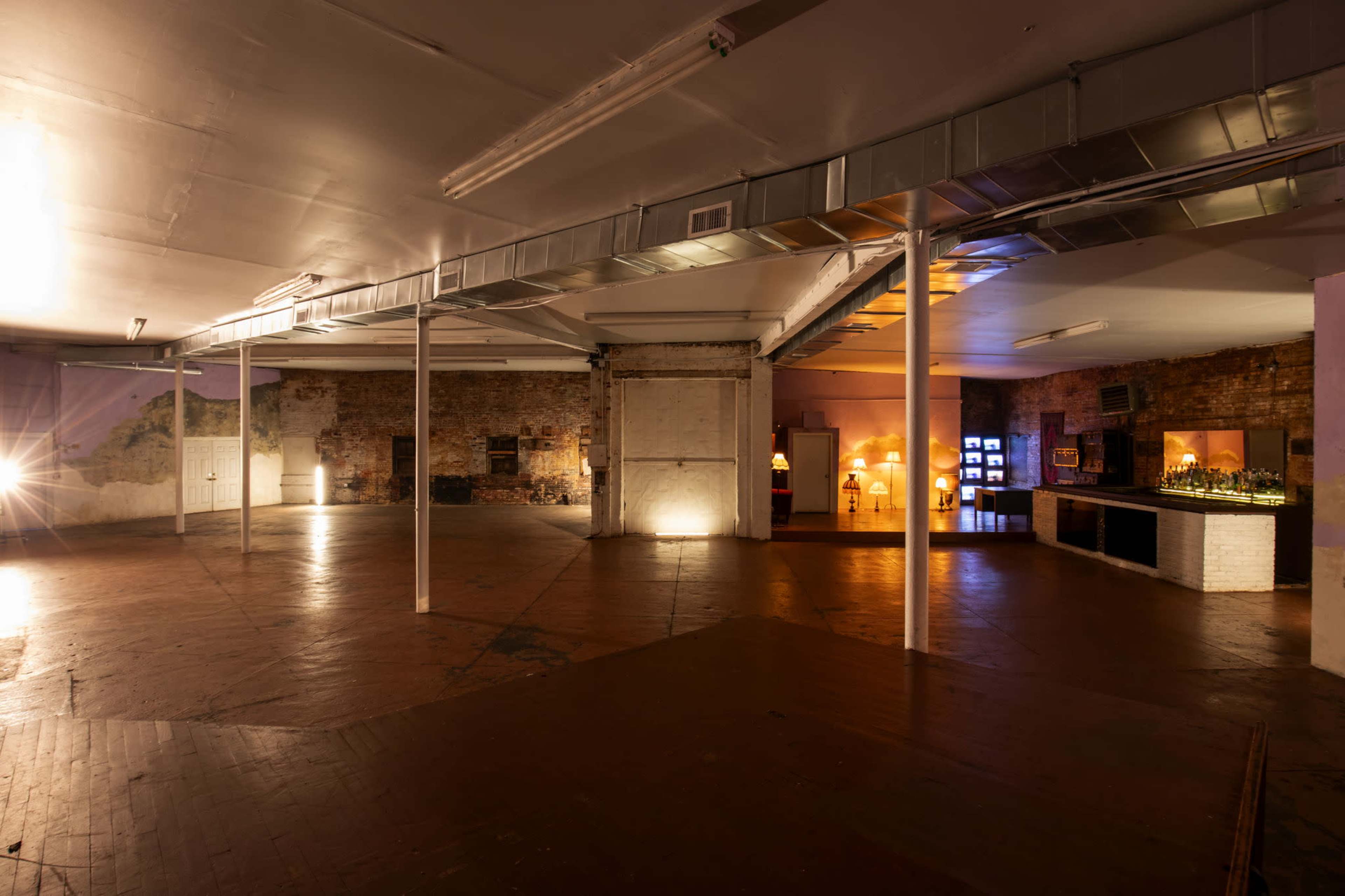 The image shows a spacious, dimly lit industrial room with exposed brick walls and a polished wooden floor, featuring a bar area and decorative lamps in the background.