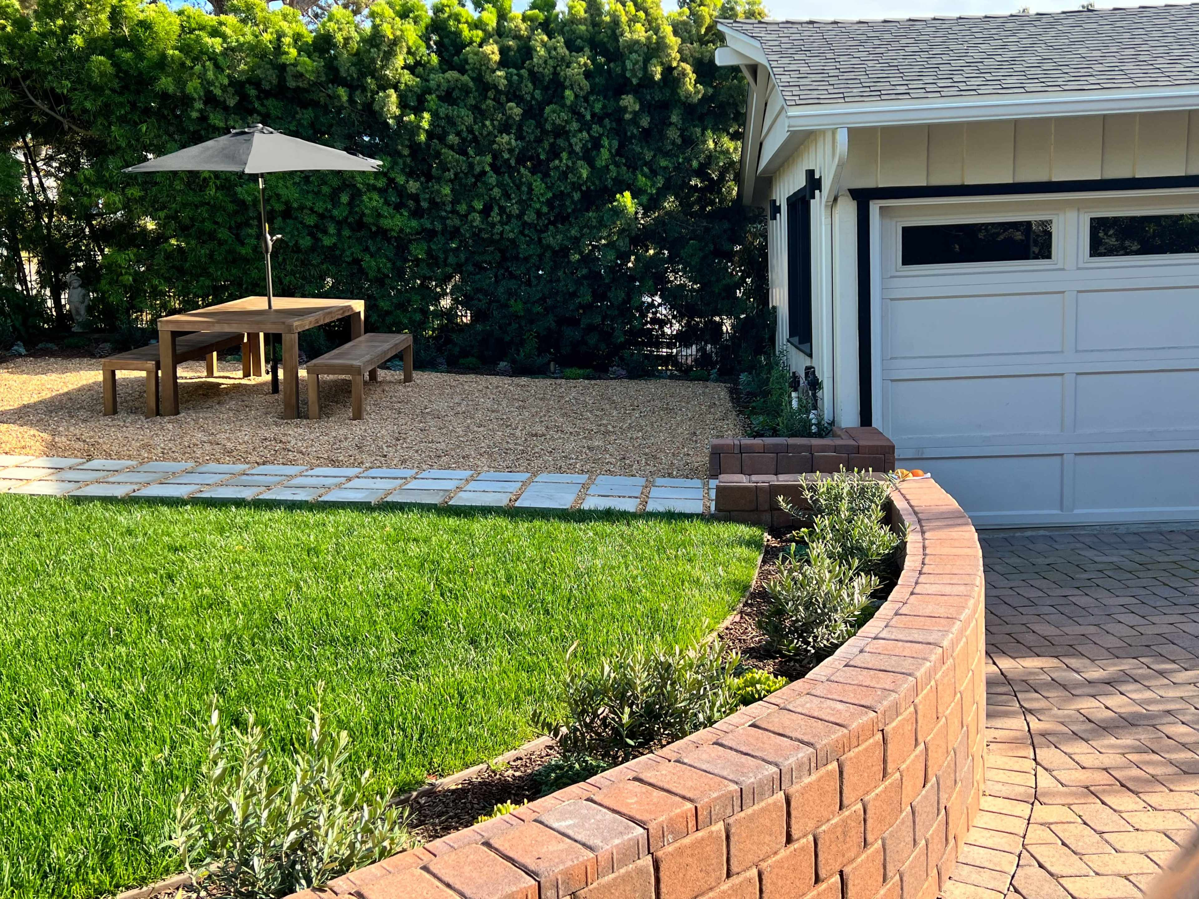 A patio area with a wooden table and benches is shaded by an umbrella, surrounded by gravel and bordered by a curved brick wall and manicured grass.