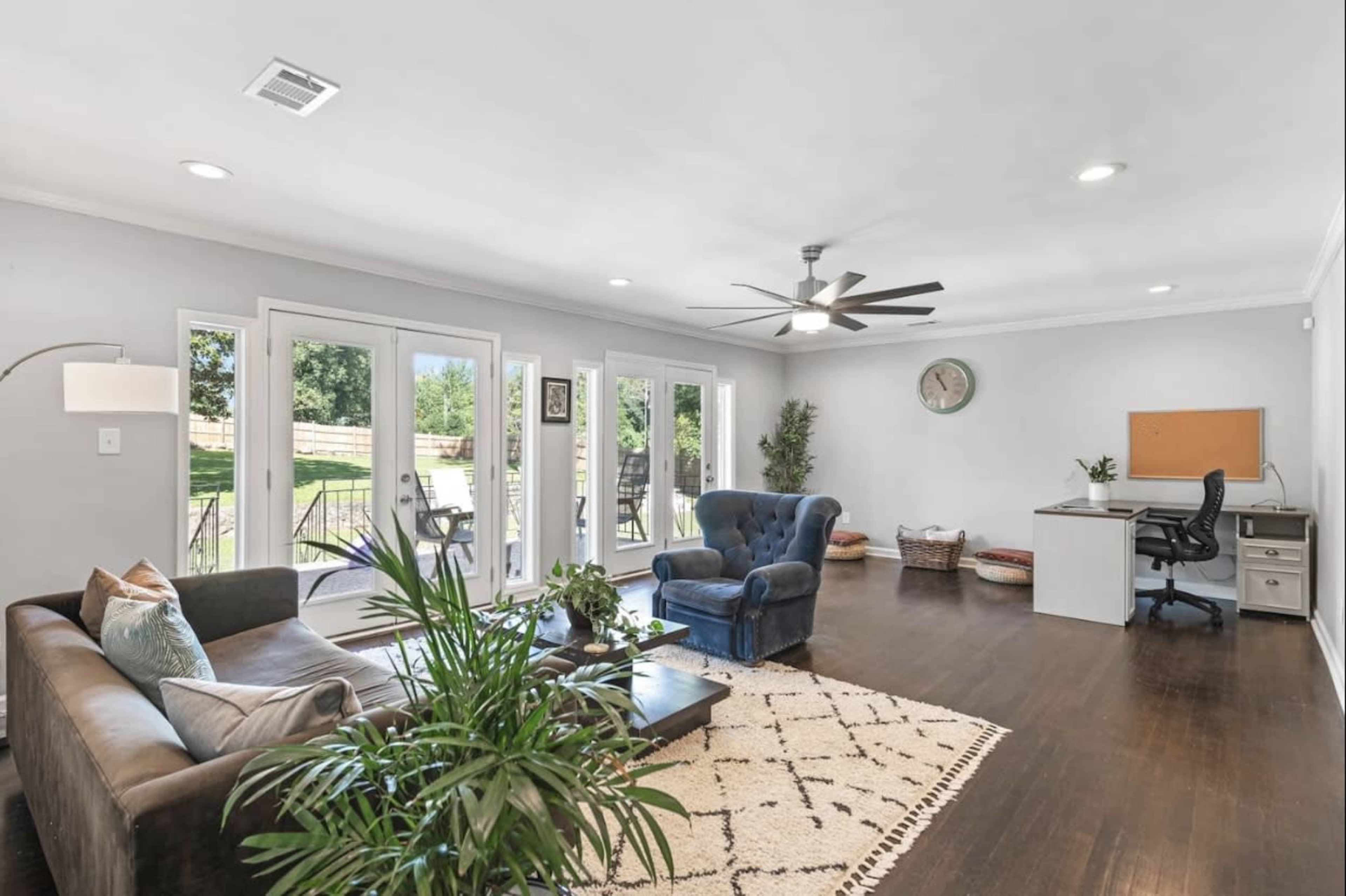 A spacious living room featuring a blue armchair, a brown sofa, a desk with a chair, and large windows that open to the outdoors.