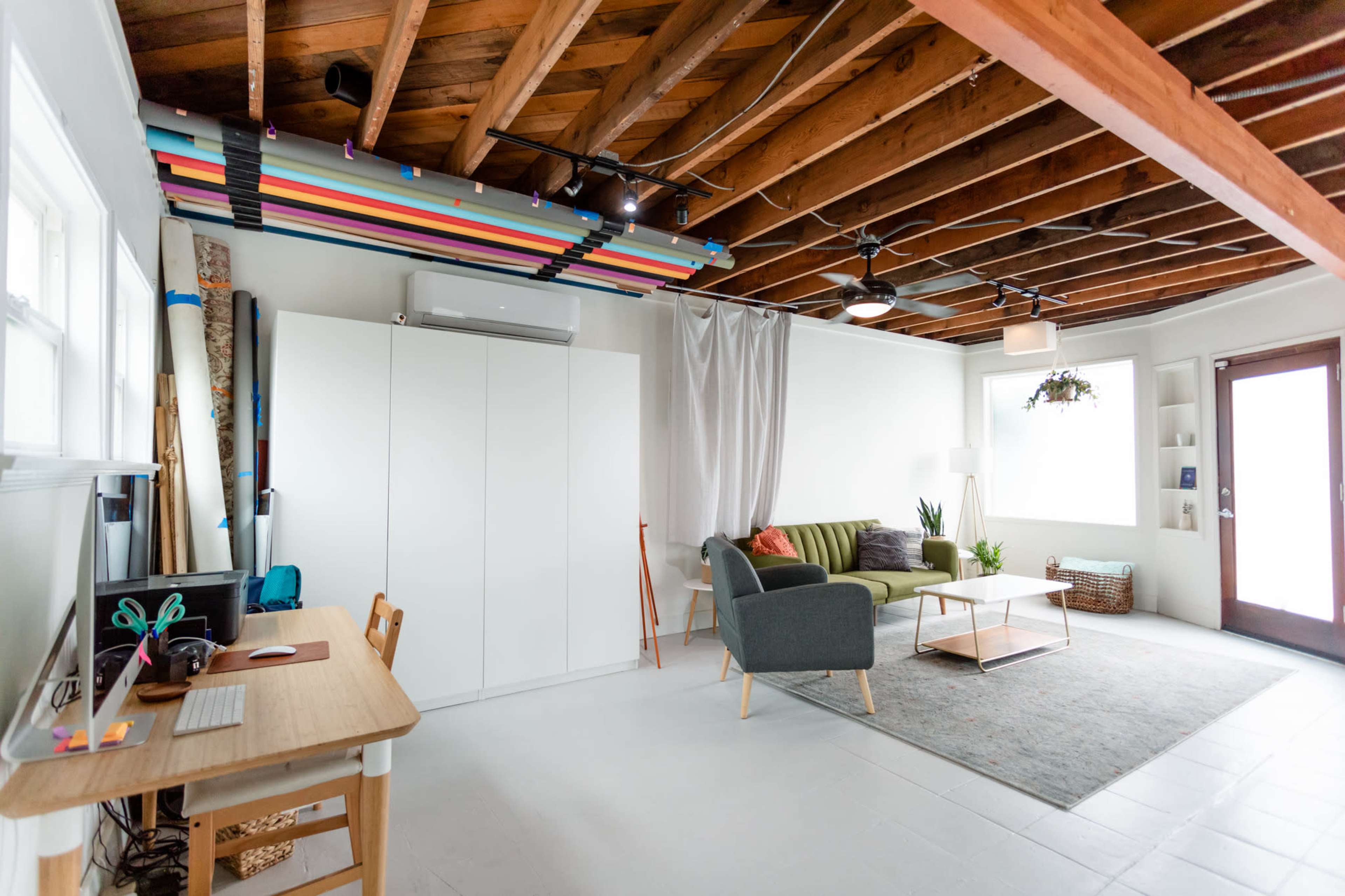 The image shows a modern, spacious living area with a green sofa, a coffee table, a desk with a computer, and an entrance door leading outside.