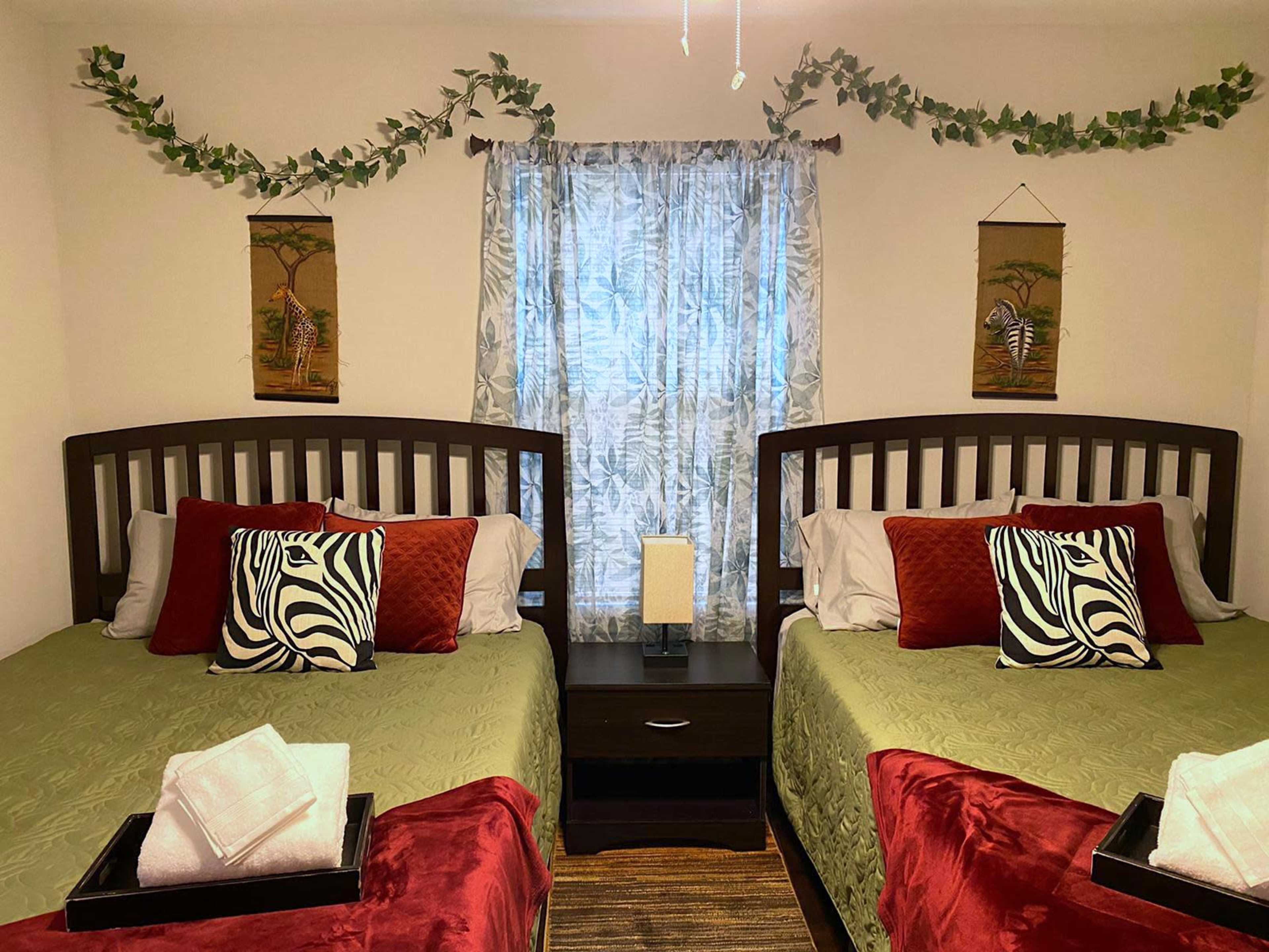 A cozy bedroom features two beds with red and green bedding, decorative zebra-print pillows, and artwork on the walls, set against a window dressed in sheer curtains.
