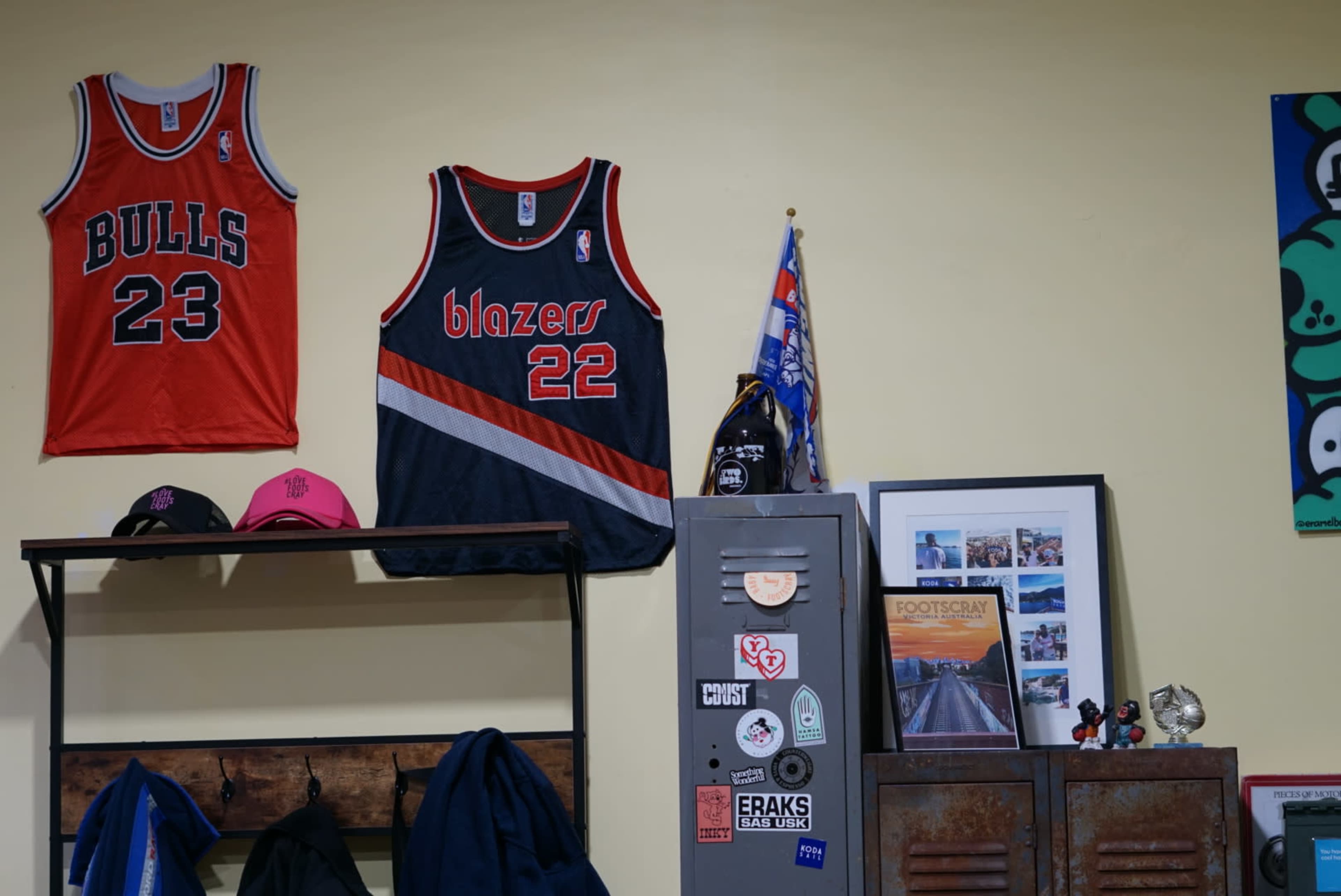 A wall with two basketball jerseys hanging above a metal locker adorned with various stickers, a framed photo, and a small flag.