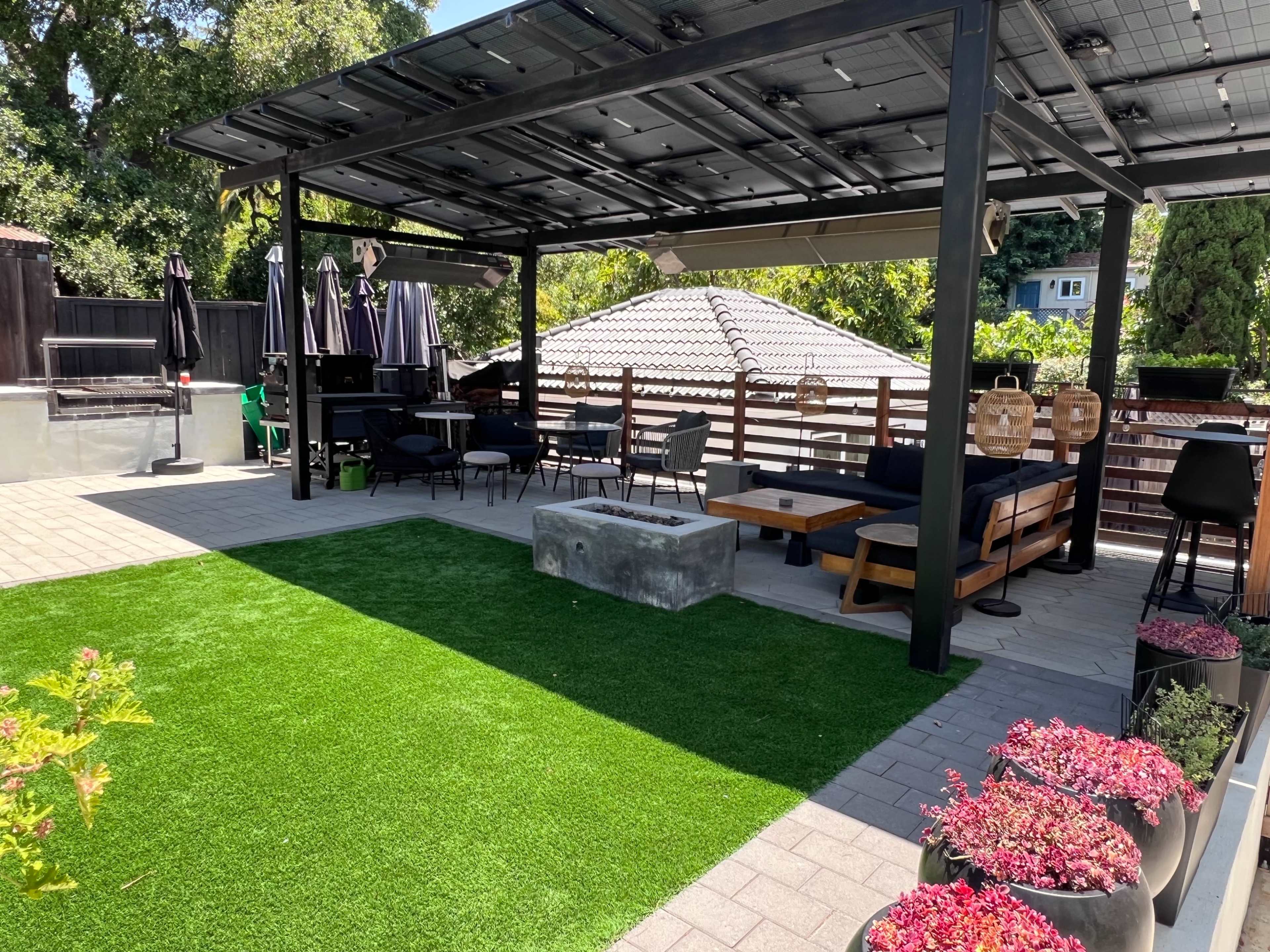 The image shows a landscaped outdoor seating area with artificial grass, a concrete table, shaded seating, and a pergola covered with solar panels.