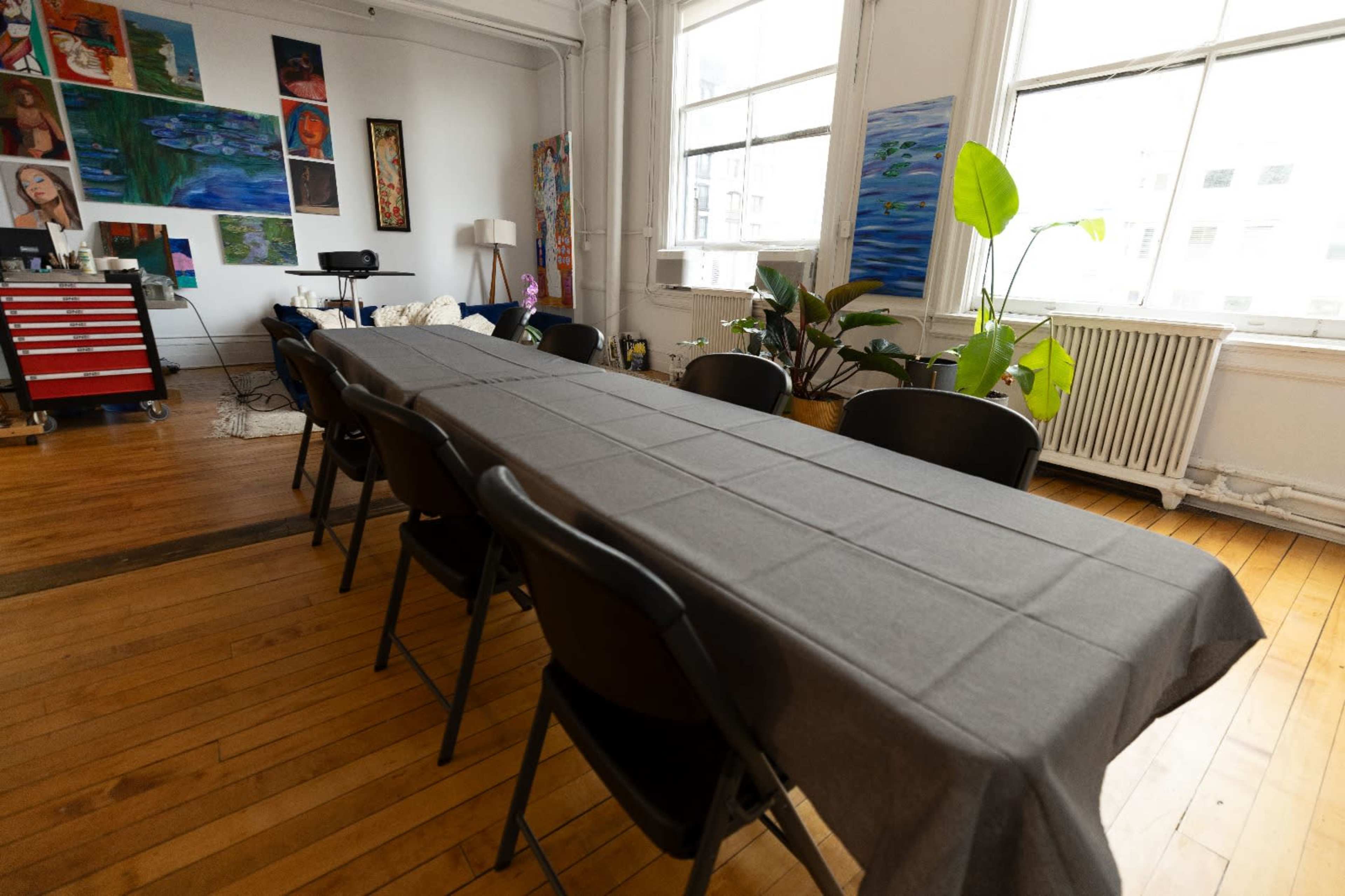 A long, rectangular table covered with a gray tablecloth, surrounded by black chairs in a bright room featuring various artworks on the walls.