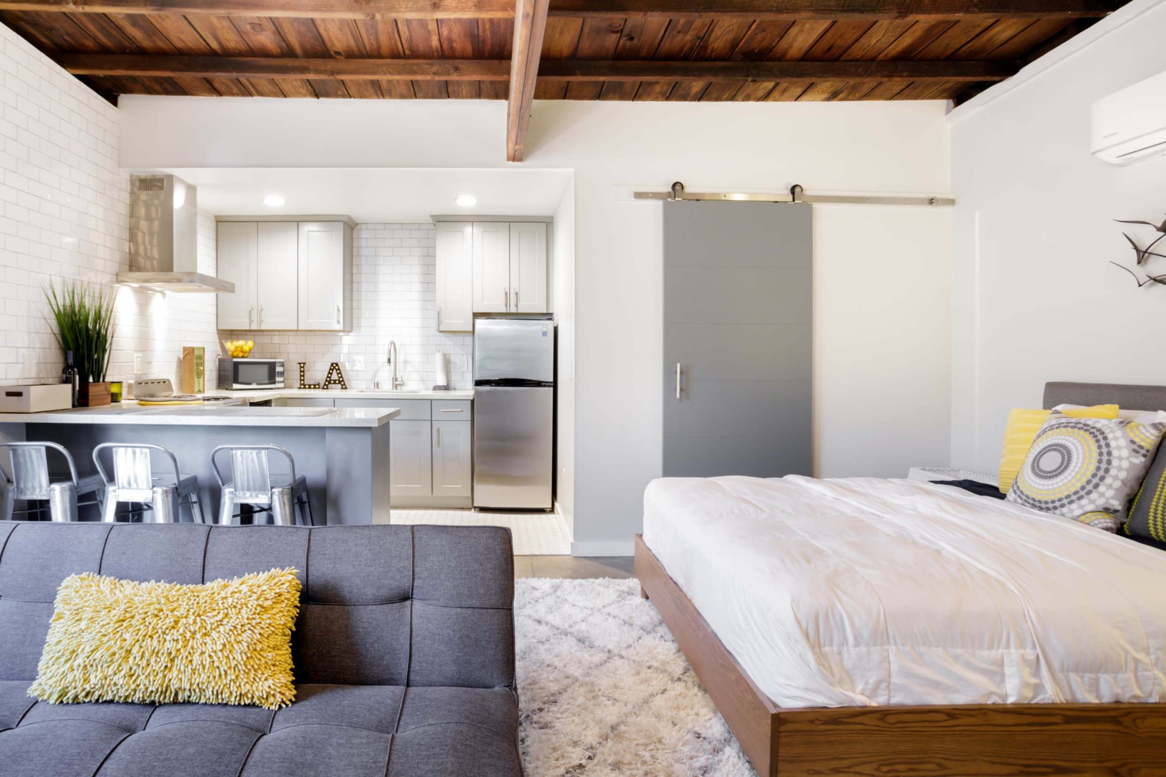 A modern studio apartment features a kitchen area with stainless steel appliances, a gray sofa with a yellow pillow, and a bed with white linens, all under exposed wooden beams.