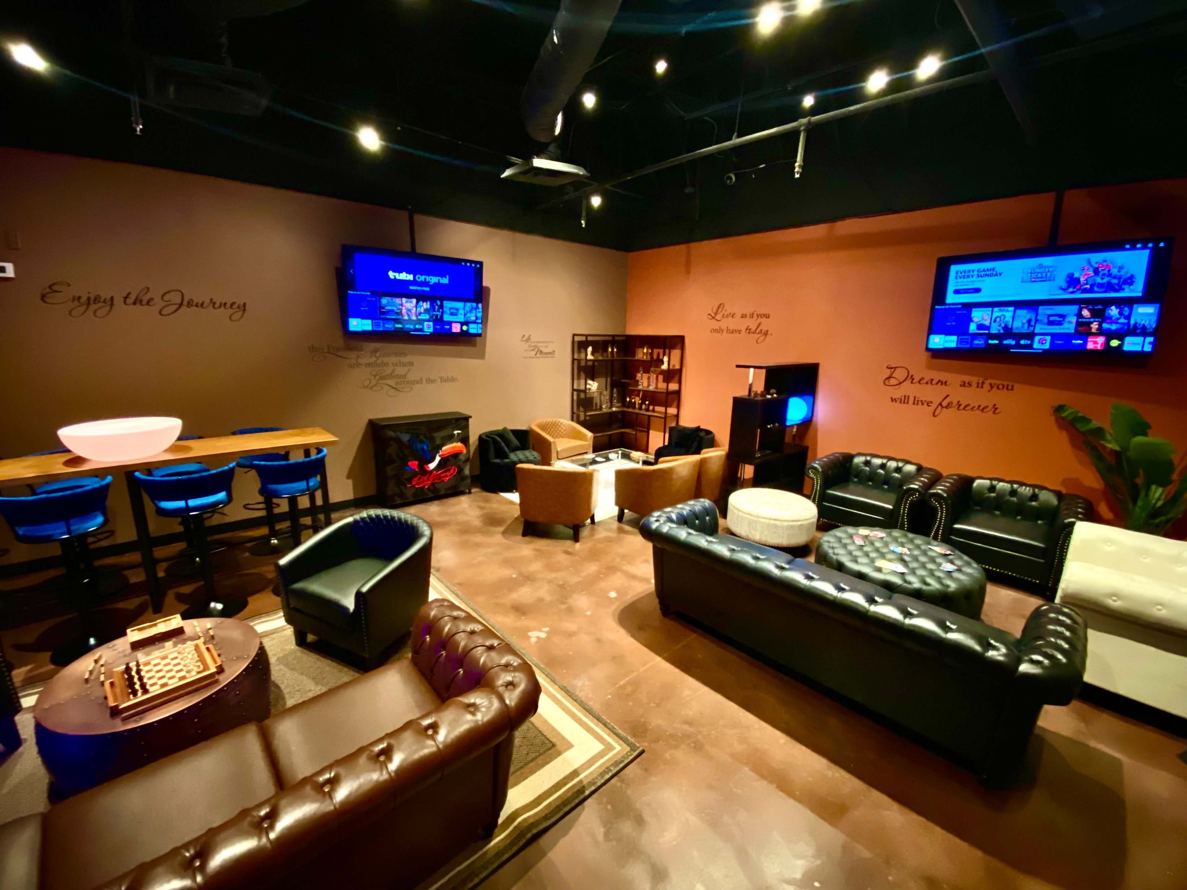 The image shows a cozy lounge area with multiple seating arrangements, two wall-mounted TVs, and decorative wall text.