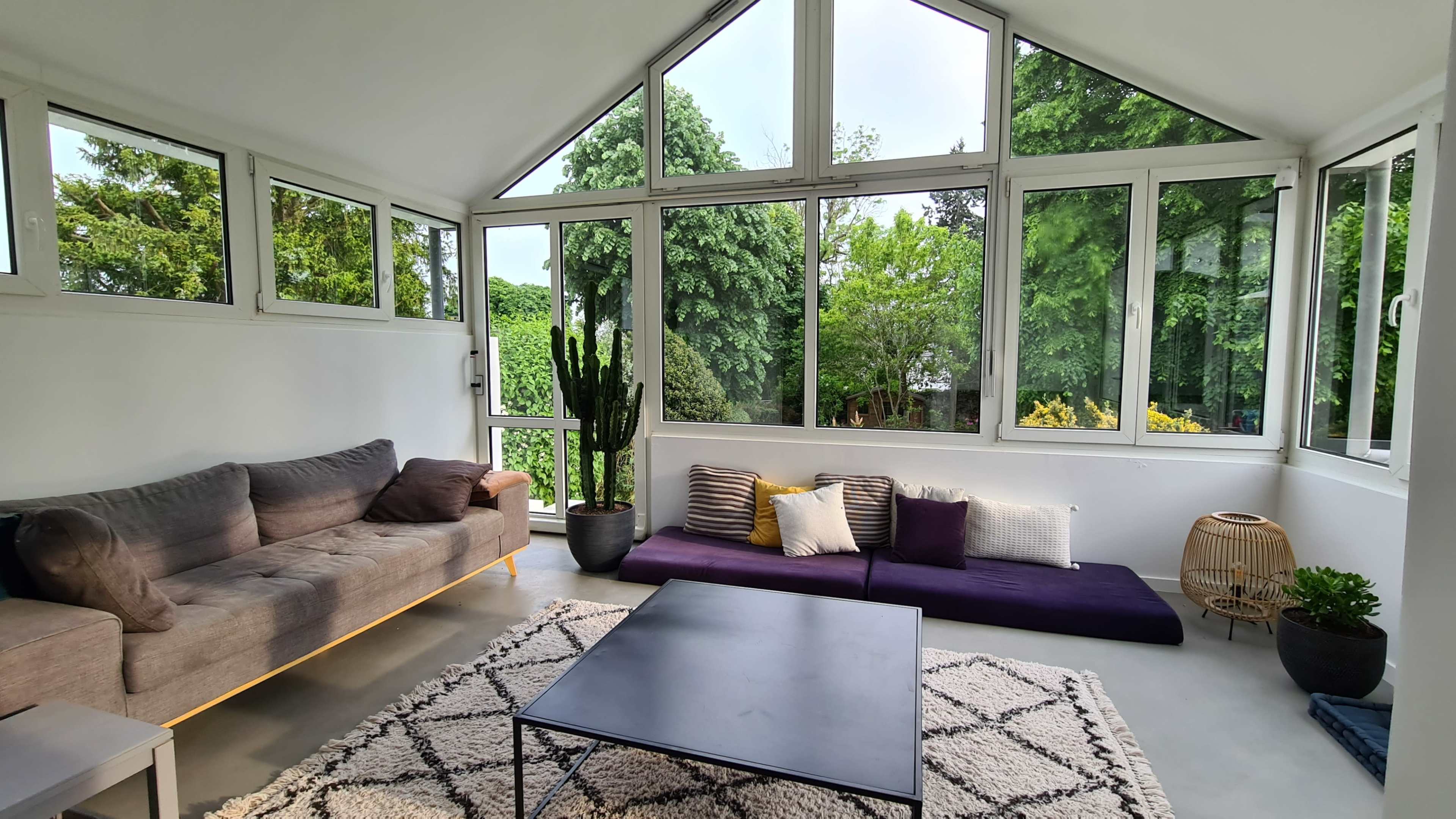 A modern living room features large windows, a gray sofa, a purple seating area, a black coffee table, and greenery visible outside.